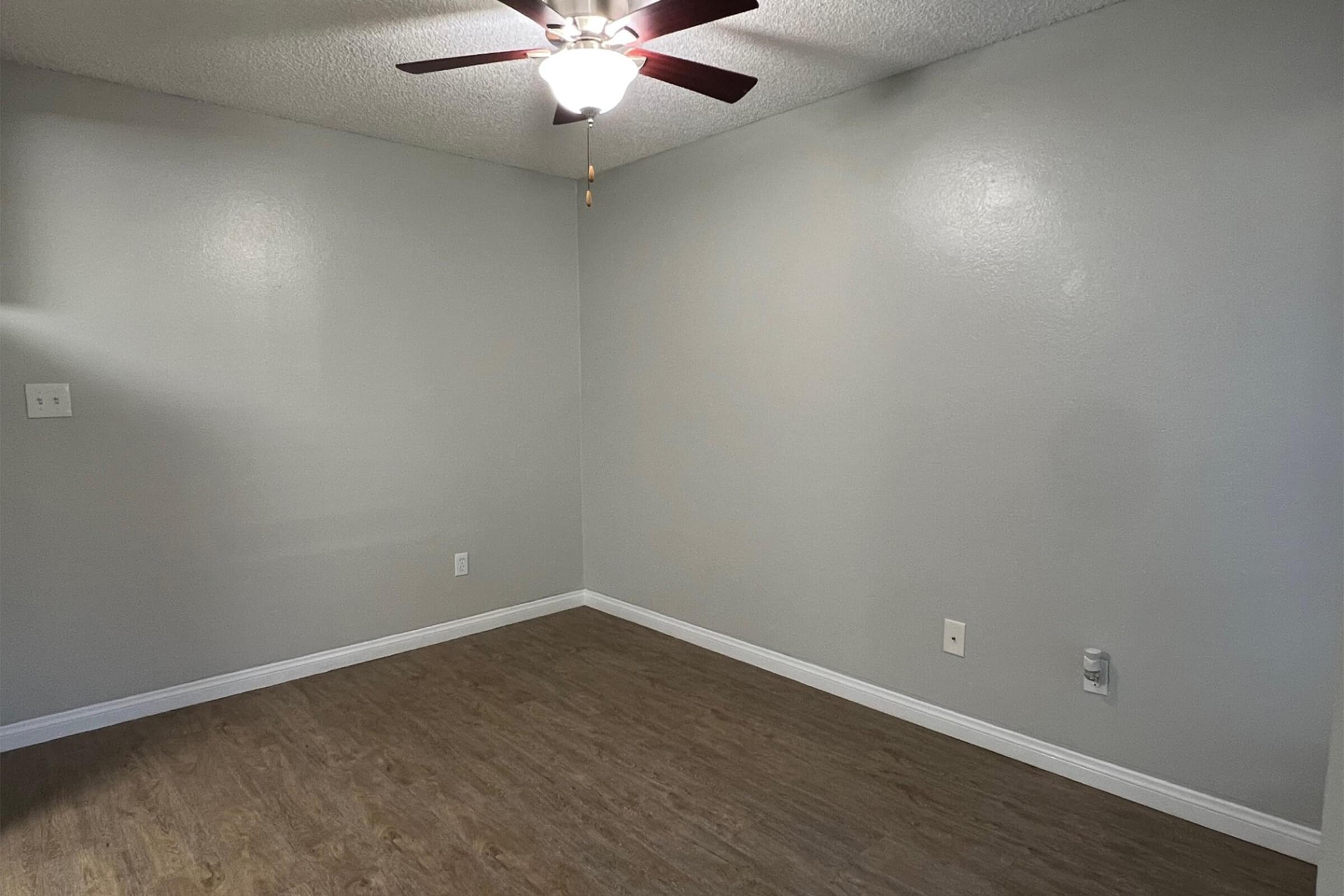 Empty room with light gray walls and a ceiling fan. The floor features wooden laminate, and there are a few electrical outlets on the walls. Natural light is minimal, creating a calm and neutral atmosphere.