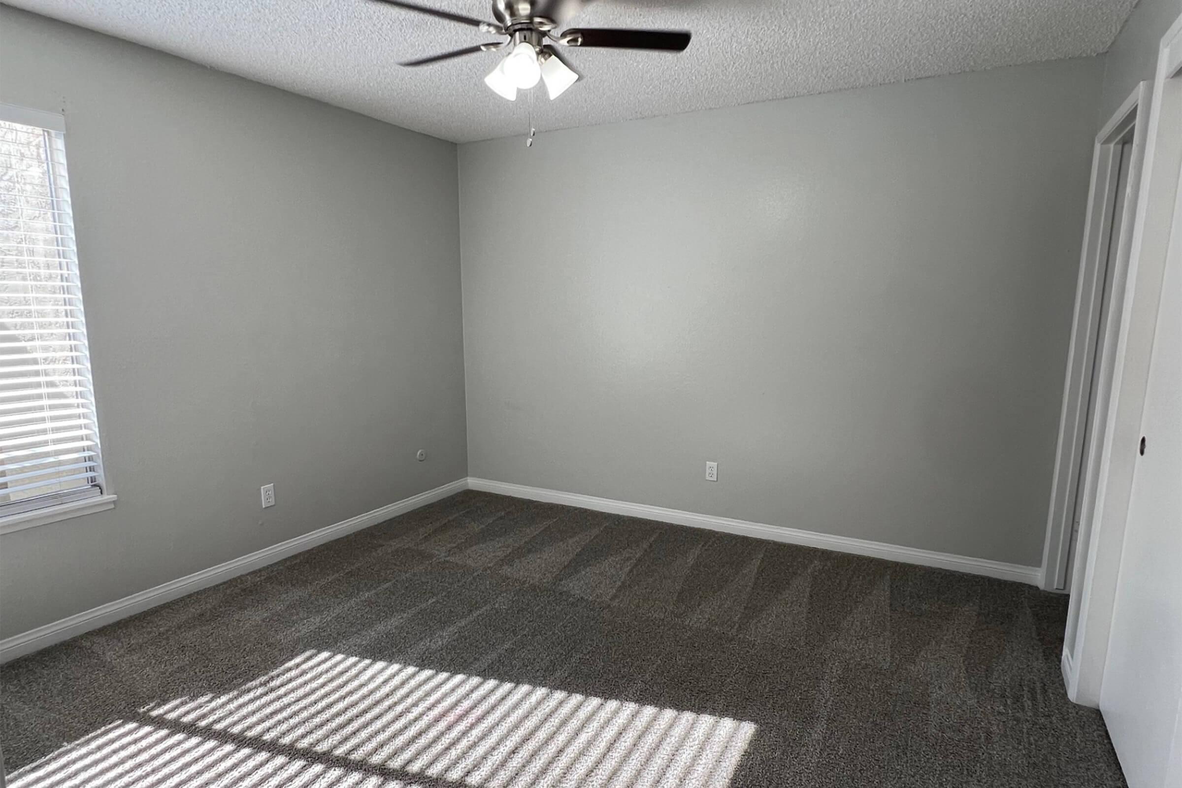 A bright and empty room with light gray walls and carpet. A ceiling fan is visible, and sunlight casts shadows on the floor from the window blinds. There is a closet door on one side and a clean, minimalist aesthetic.