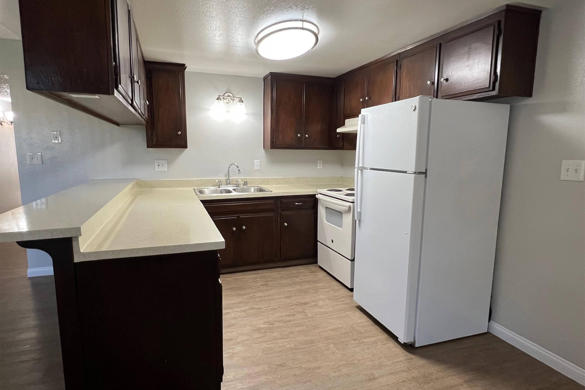 A modern kitchen featuring dark wooden cabinets, light countertops, a white refrigerator, and a stove. The room is well-lit with a ceiling light fixture, showcasing an open layout. The floor has light wood-like laminate, contributing to a clean and spacious appearance.