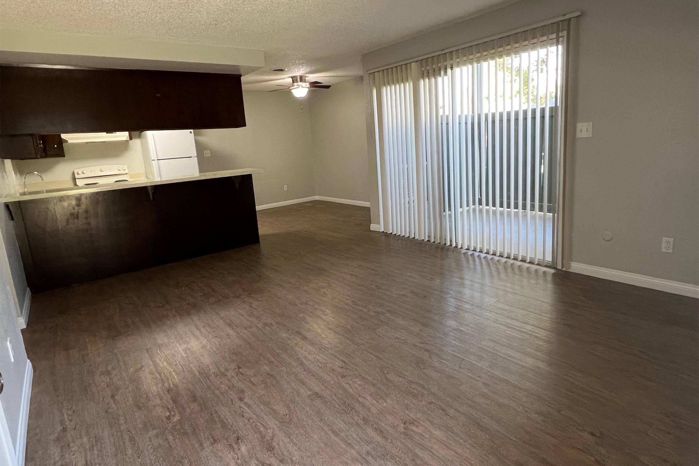 Spacious living area with light wood flooring, a ceiling fan, and large windows with vertical blinds. An open kitchen features dark cabinets and appliances, including a white refrigerator and stove. The space is bright and airy, with a view of an outdoor area through the sliding glass door.