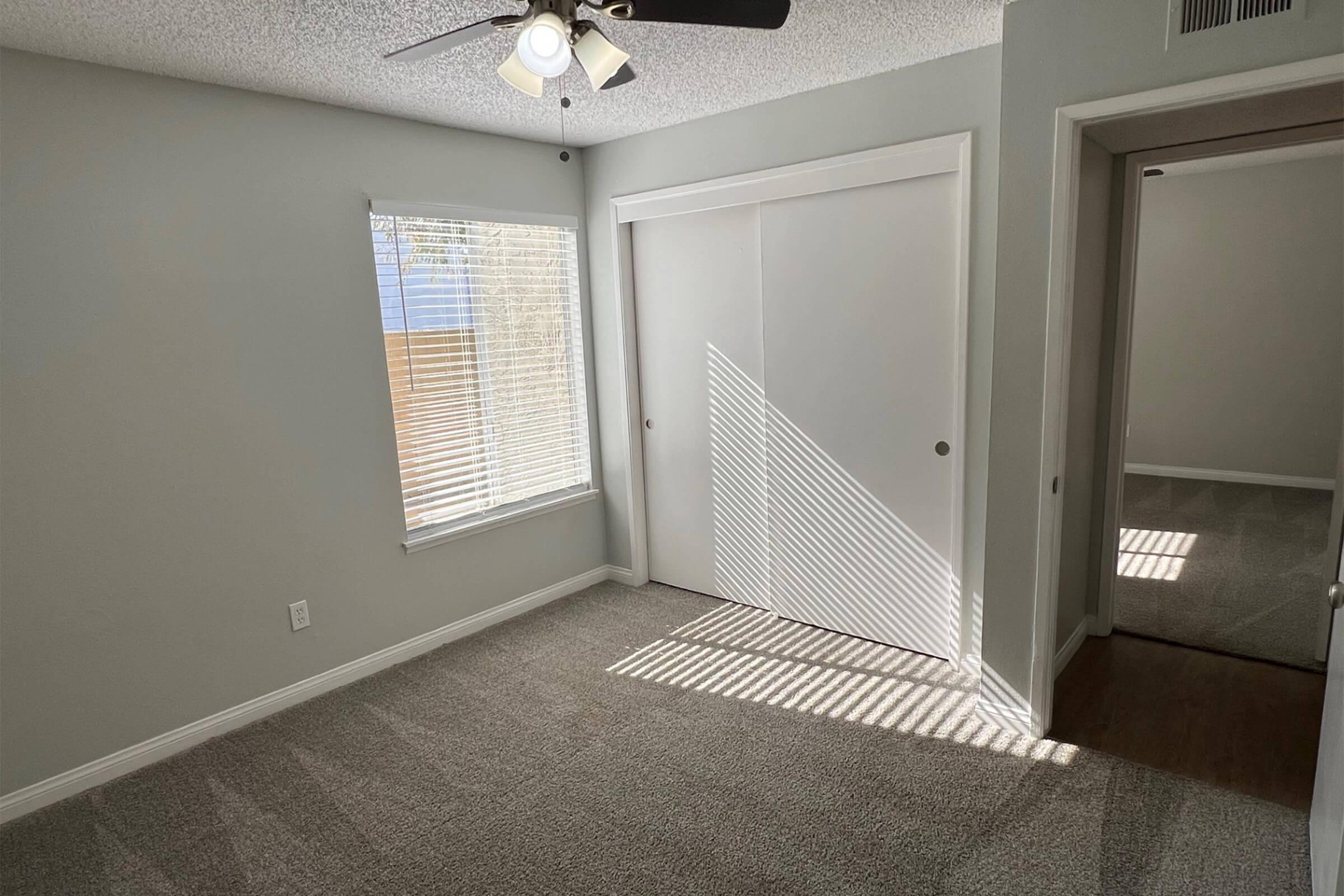 A well-lit room with gray walls and carpet, featuring a window with blinds casting shadows on the floor. There's a ceiling fan and a closet with sliding doors. The adjacent doorway leads to a small hall or another room, enhancing the spacious feel.