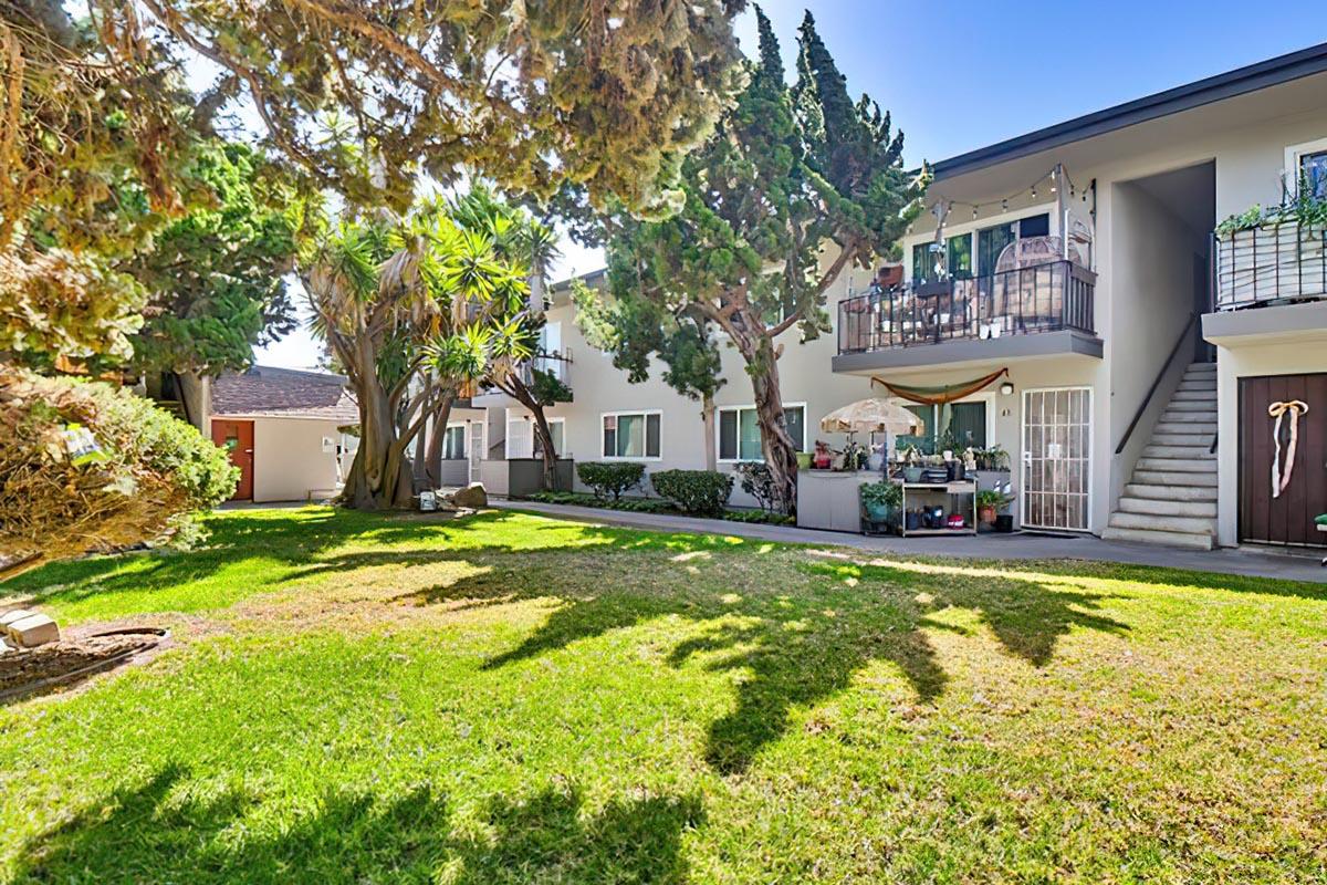 A serene outdoor area featuring lush green grass and tall trees surrounding an apartment complex. The building has balconies adorned with plants and decorations. Sunlight casts shadows on the lawn, creating a peaceful atmosphere. In the background, there's a staircase leading to the upper level of the apartments.