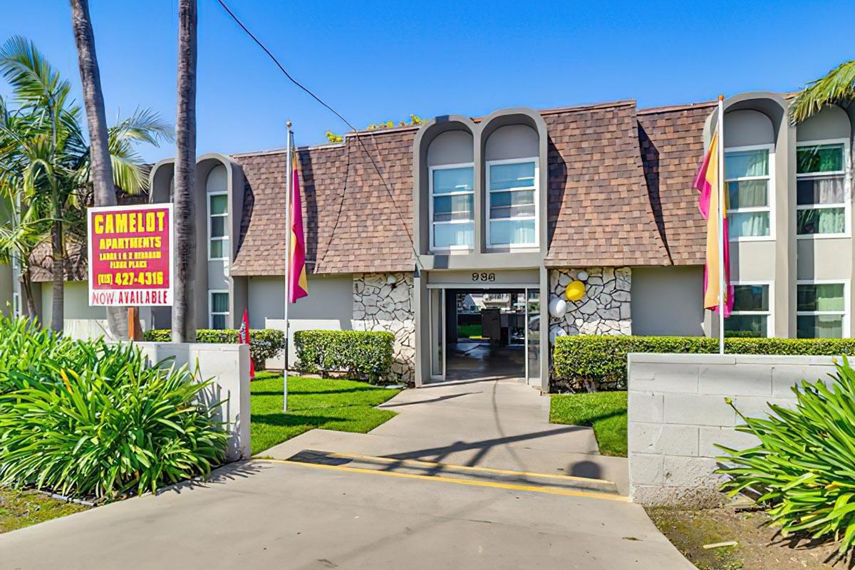 Front view of Camelot Apartments featuring a stone and stucco exterior, large windows, and a sloped roof. The entrance is flanked by palm trees and colorful flags. A sign near the entrance advertises availability, with the phone number prominently displayed. The area is well-maintained with green landscaping.