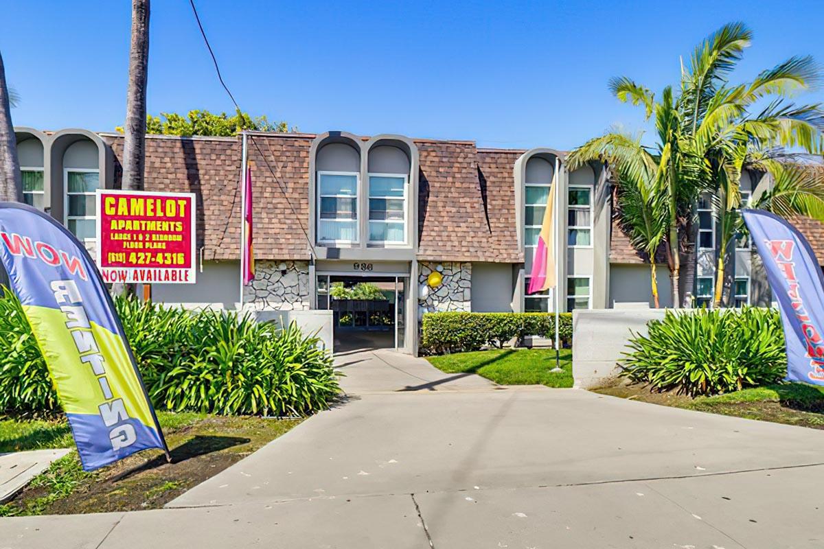 Exterior view of Camelot Apartments featuring a modern building with a distinctive shingled roof. The entrance is flanked by colorful flags and palm trees, with greenery around the property. A sign indicates that rental units are available, along with a phone number for inquiries. Clear blue sky in the background.