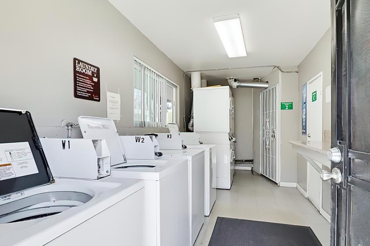 A clean and well-lit laundry room featuring several washing machines labeled W1, W2, and W3. A dryer is also visible, along with informational signage on the wall. The space has a neutral-colored interior with a door leading to another area. A large window provides natural light.