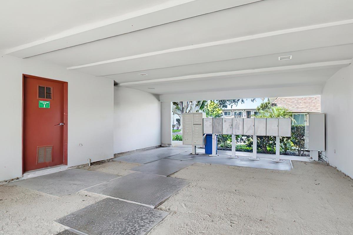 An empty interior space featuring a door on the left, a concrete floor, and a row of mailbox compartments against the wall. Natural light illuminates the area, and a view of greenery is visible through an opening. The walls are painted white, and the overall atmosphere is minimalist and modern.