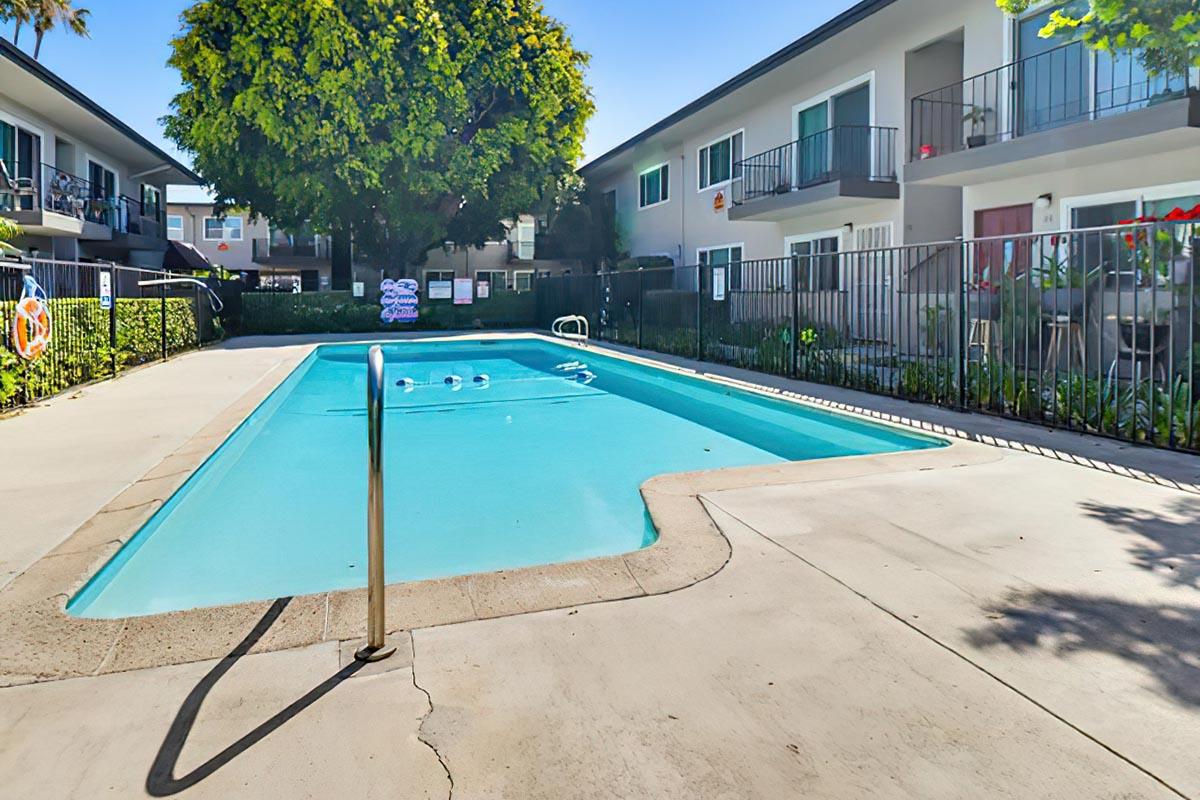 A rectangular swimming pool surrounded by a concrete deck, located in a courtyard area with two-story apartment buildings in the background. Lush greenery and some decorative potted plants are visible. The sky is clear and sunny, enhancing the inviting atmosphere of the pool area.