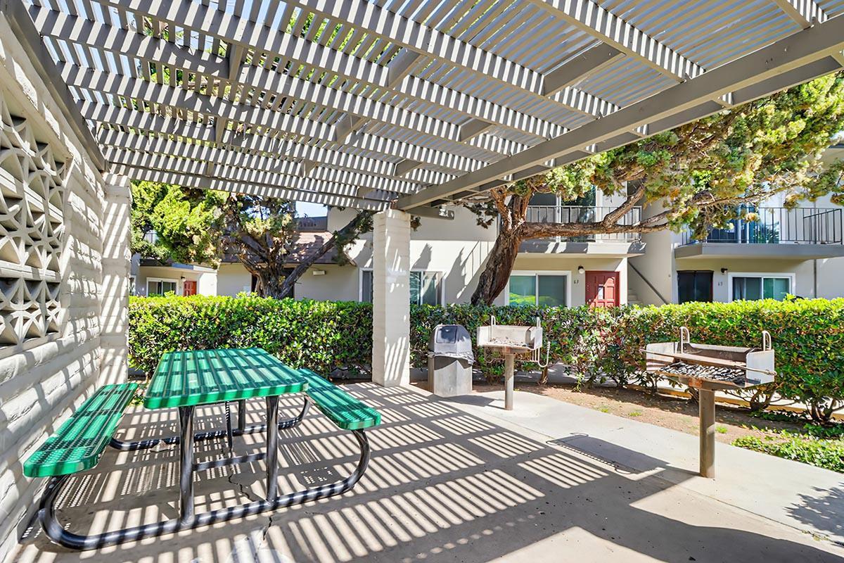 Outdoor picnic area featuring a green picnic table under a striped pergola, surrounded by neatly trimmed hedges. In the background, there are residential buildings with balconies. Two barbecue grills are visible: one is a stationary grill and the other is a portable model. The setting is bright and inviting.