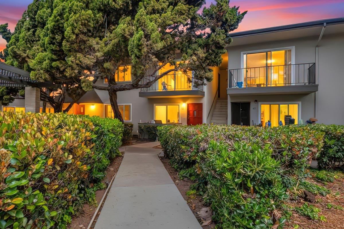 A pathway leading to a two-story building surrounded by greenery. The building features balconies with warm lights illuminating the windows, set against a colorful sunset sky. The scene conveys a tranquil and inviting atmosphere.