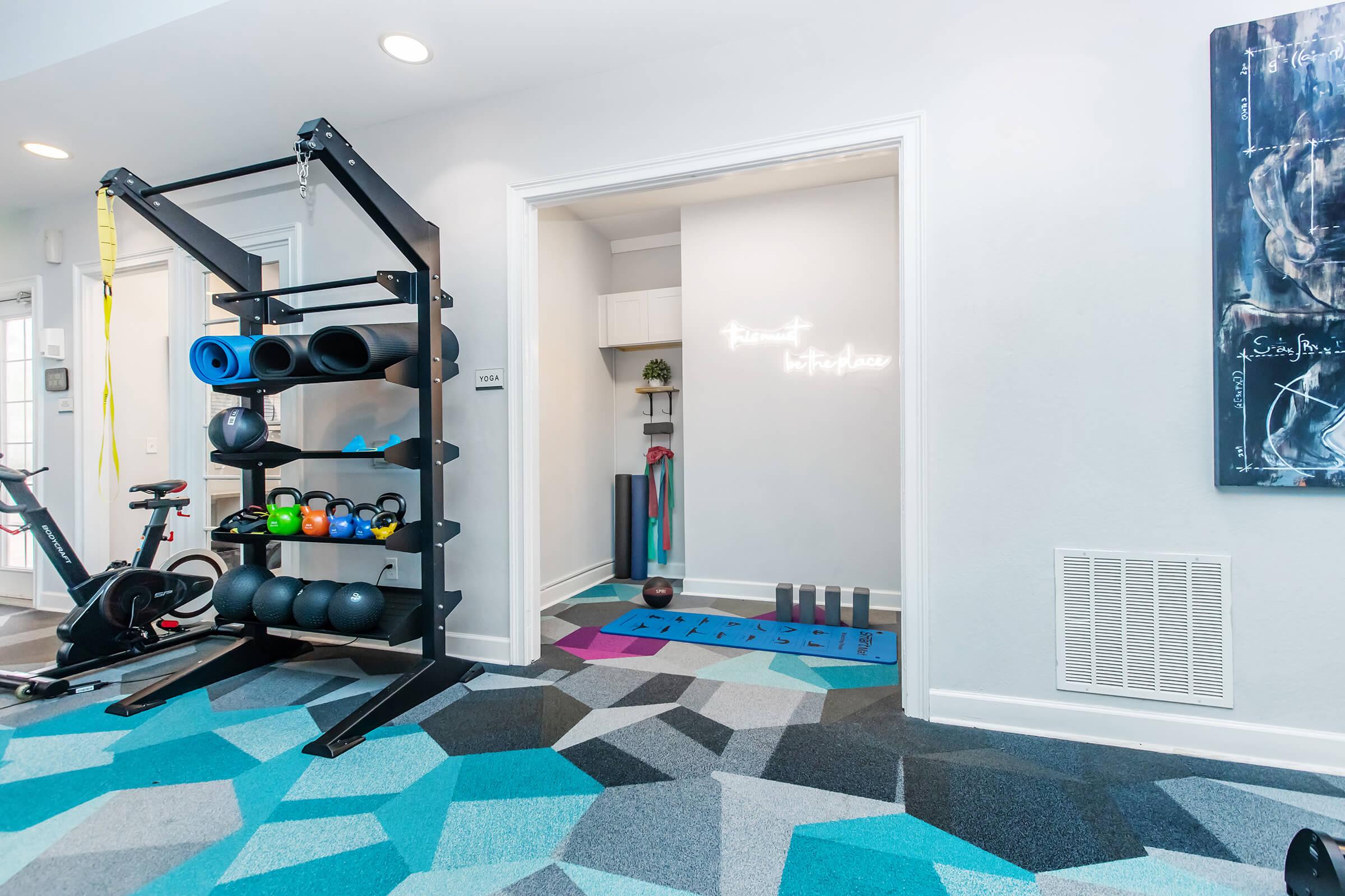 A modern workout space featuring a black weight rack with colorful dumbbells and exercise mats, a stationary bike, and a colorful geometric carpet. The wall has a motivational quote illuminated in light. A doorway leads to a small area with fitness equipment, adding to the functional design of the gym.