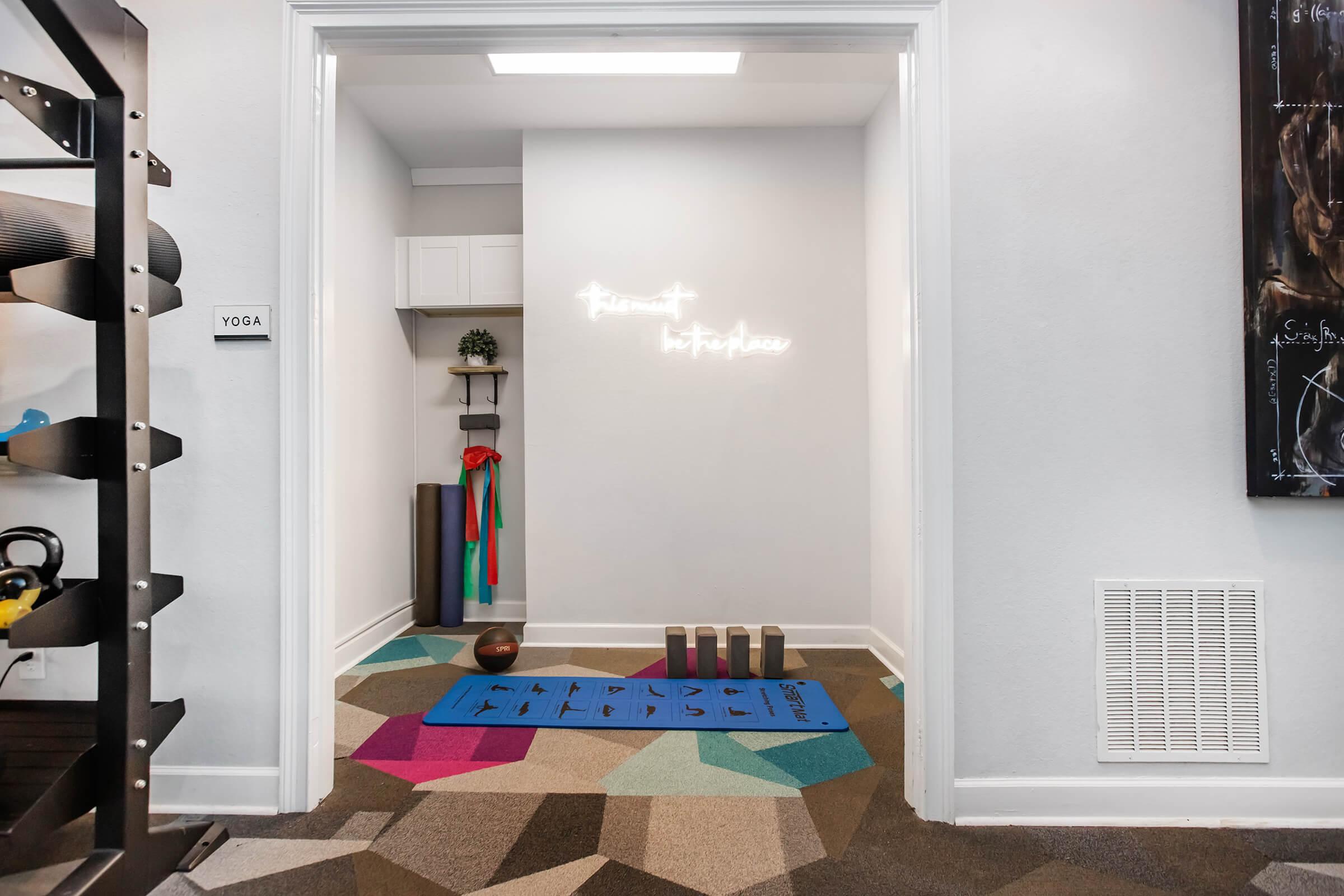 Yoga corner in a bright room featuring a blue yoga mat, colorful resistance bands, and a small shelf with a plant. The wall displays a glowing sign that reads "Present in the moment." A variety of exercise equipment is visible, along with a door marked "YOGA" on the left side.