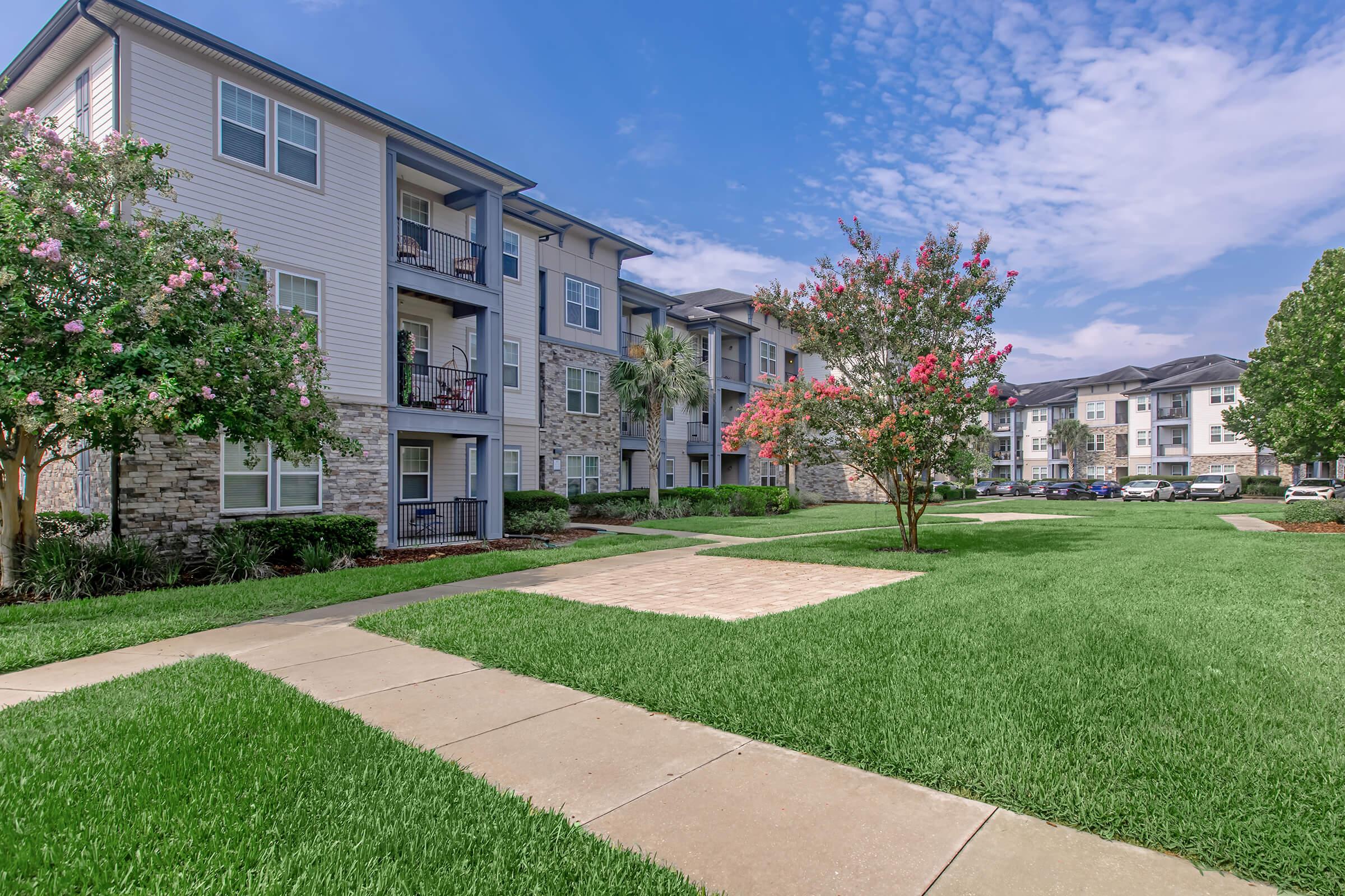 A well-maintained apartment complex featuring three-story buildings with balconies, surrounded by lush green lawns and flowering trees. The sky is partly cloudy, and there is a paved walkway leading through the grass. Parking spaces are visible in the background.