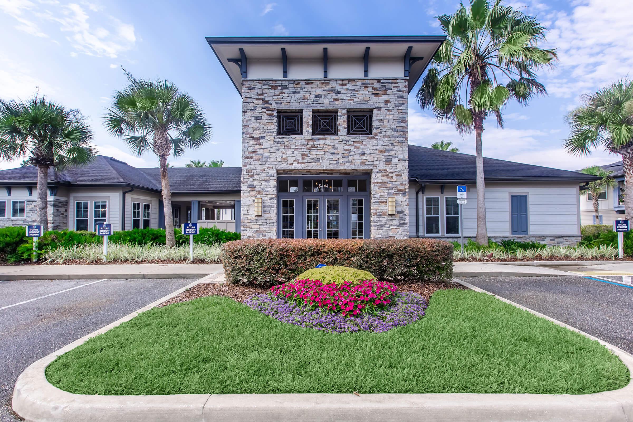 A modern building with a stone facade and large windows surrounded by palm trees. In the foreground, a landscaped area features vibrant flowers and lush green grass, creating an inviting entrance. The sky is clear with some clouds, suggesting a pleasant day.