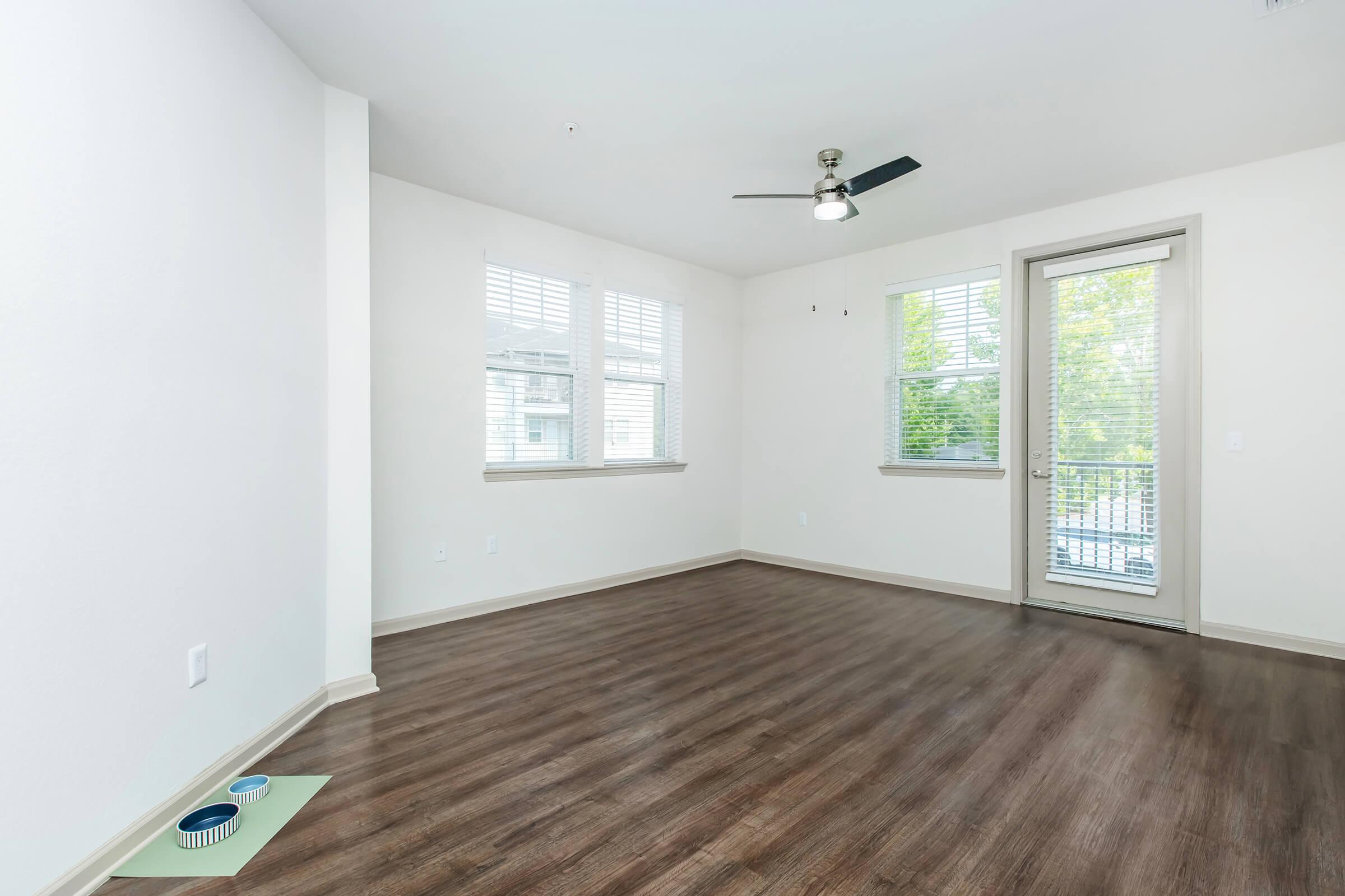 A spacious, empty living room with light-colored walls and wooden flooring. It features a ceiling fan, two large windows letting in natural light, and a sliding glass door leading to a balcony. A small pet bowl is placed on the floor near the wall.