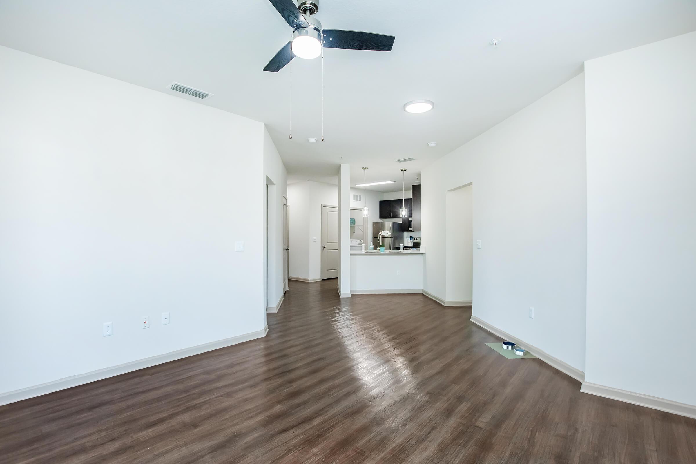 A spacious, well-lit interior of an apartment featuring hardwood flooring, a ceiling fan, and an open layout. The kitchen is visible at the end of the room, with doorways leading to other areas of the apartment. The walls are painted in a neutral color, enhancing the bright and airy atmosphere.