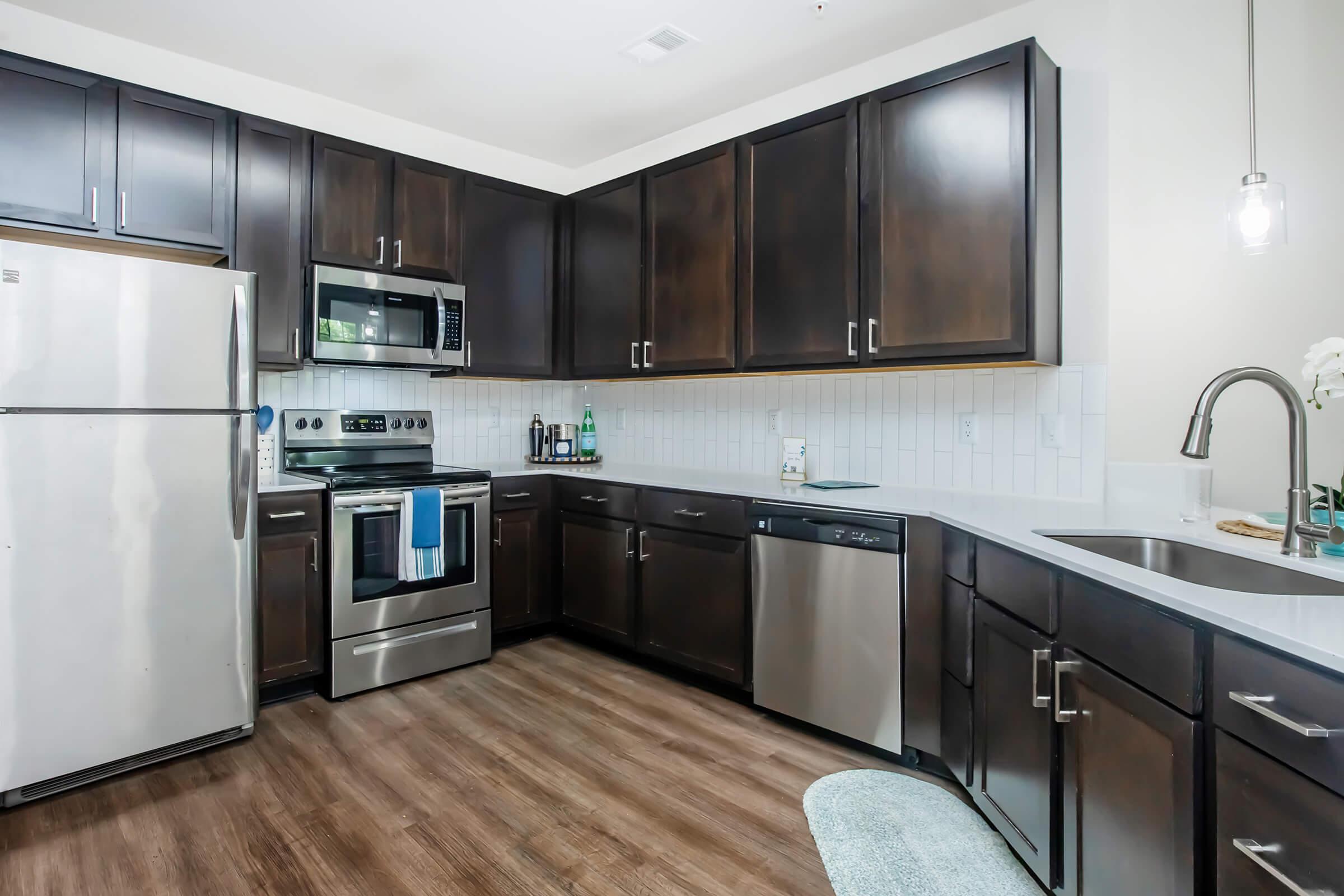 Modern kitchen featuring dark wood cabinets, stainless steel appliances including a refrigerator, oven, and microwave. Countertop with a shiny surface and a sink. A blue dish towel hangs on the oven. The flooring is wood-like, and there is a small light fixture above the countertop.