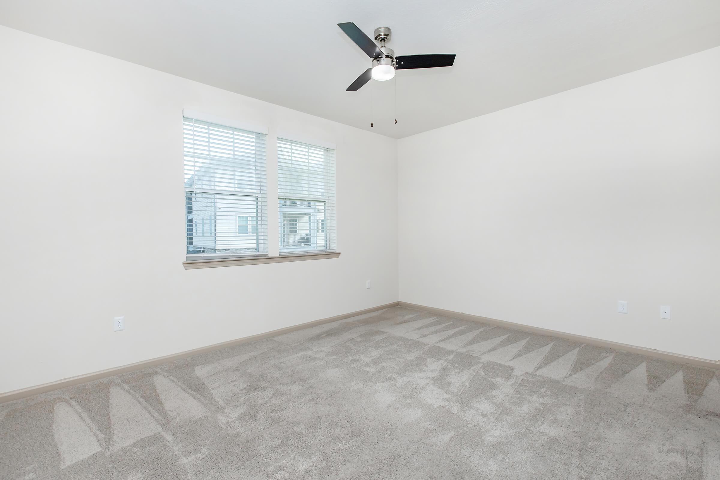 Empty room with light-colored walls and a ceiling fan. A large window with horizontal blinds allows natural light to fill the space. The floor is covered with light gray carpet, and there are no furnishings. The overall atmosphere is bright and airy, suggesting potential for personalization.