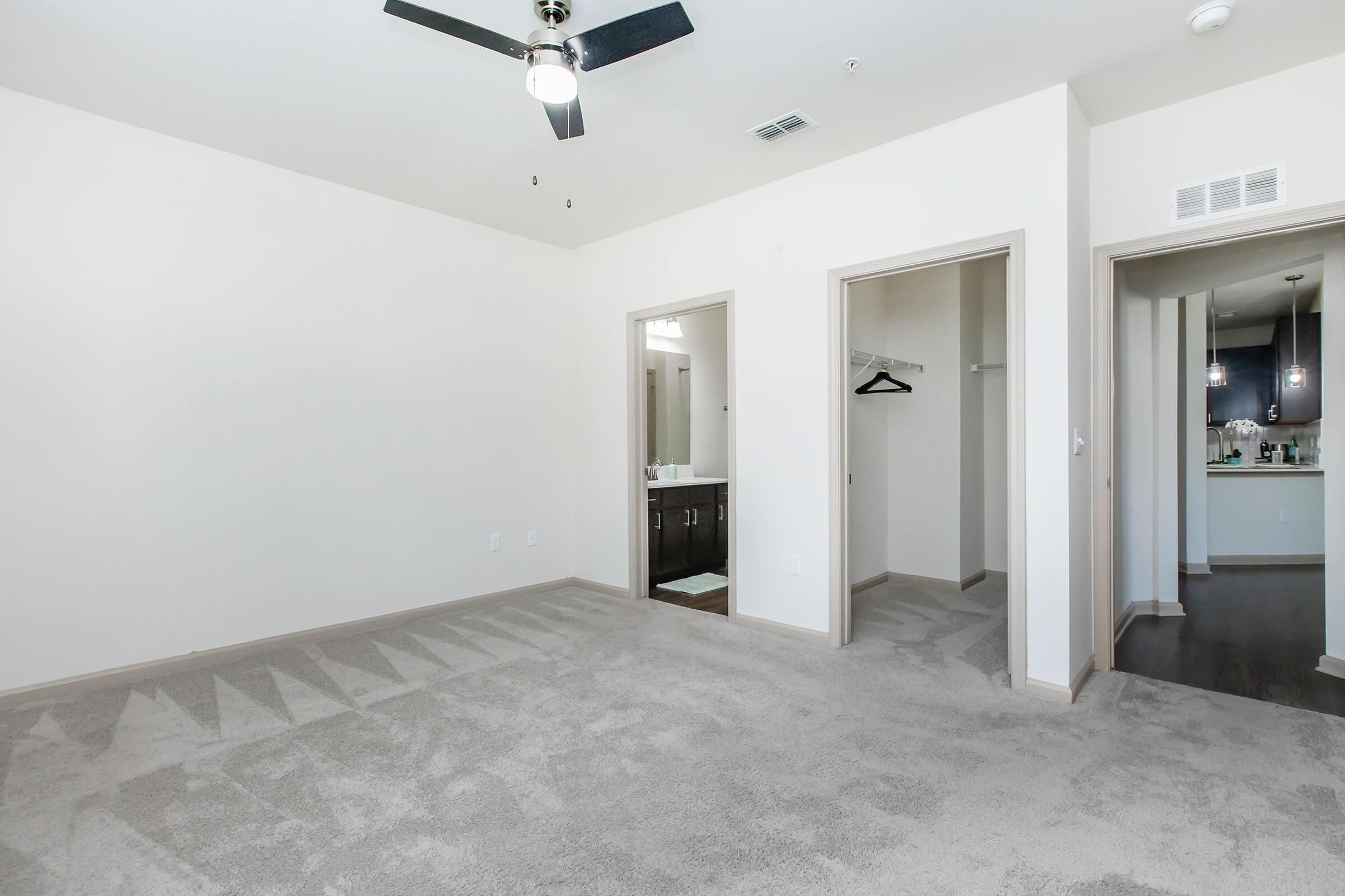 A spacious room with light-colored walls and gray carpet. There are two doorways leading to adjacent areas, one likely a bathroom with a vanity and the other a closet. A ceiling fan is visible, and the room appears bright with natural light coming from an unseen source.