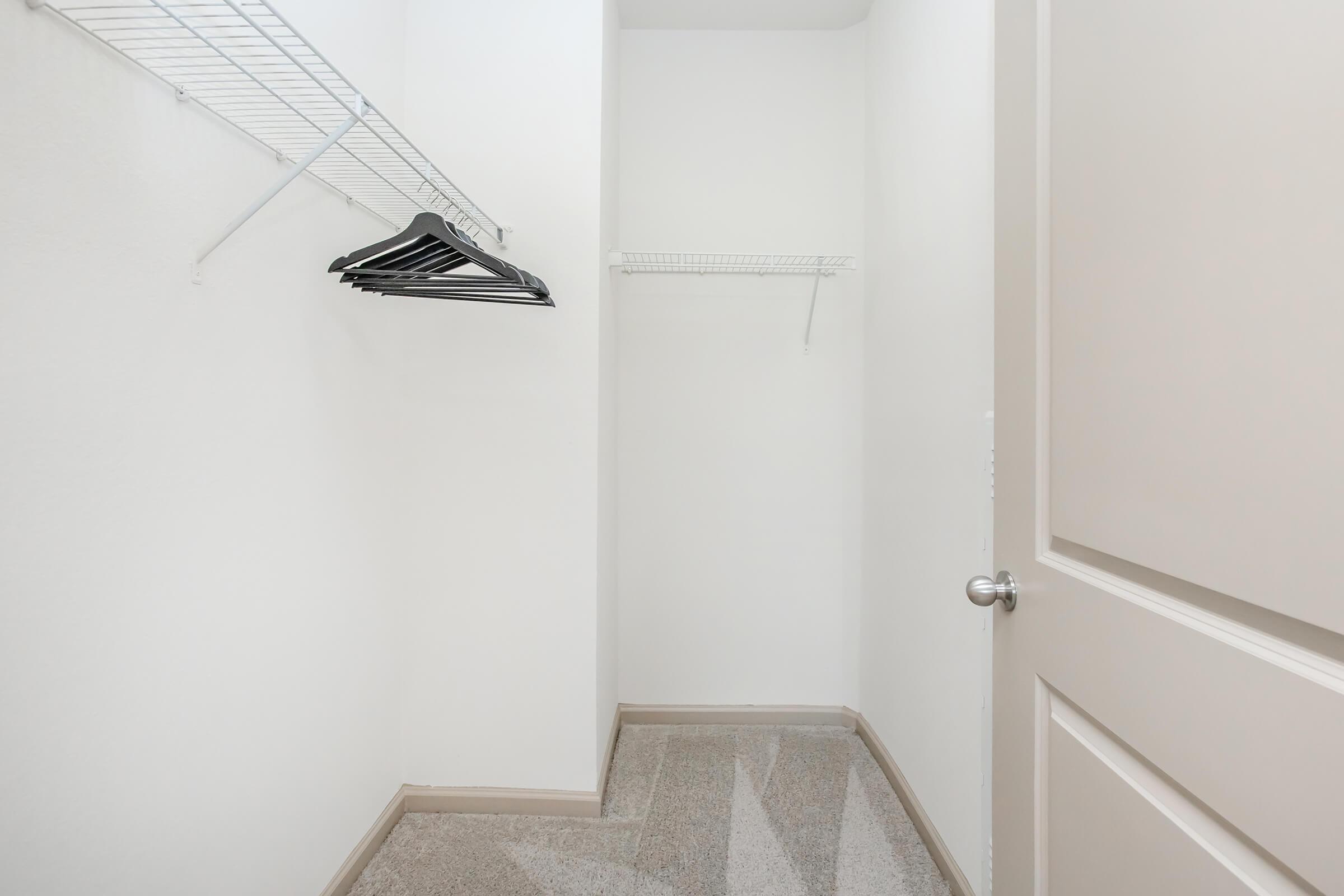 A small, empty closet with two wire shelves. One shelf has black hangers, while the other side is vacant. The walls are painted white, and there's a light-colored floor. The closet door is partially visible on the right.