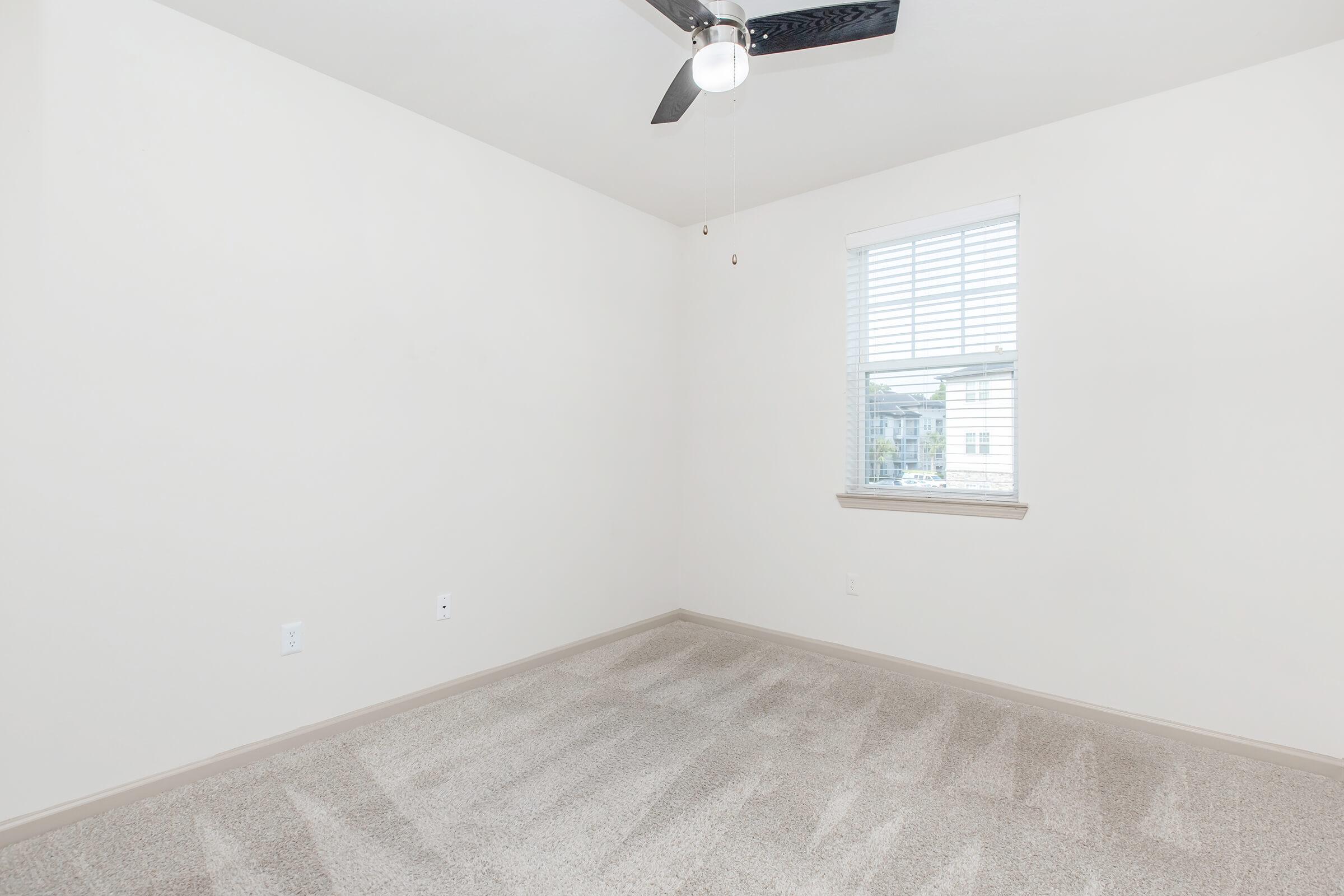 A clean, empty room with light-colored walls and beige carpet. It features a ceiling fan and a window with horizontal blinds, allowing natural light to enter. The space appears bright and minimalist, suitable for various uses, such as a bedroom or office.