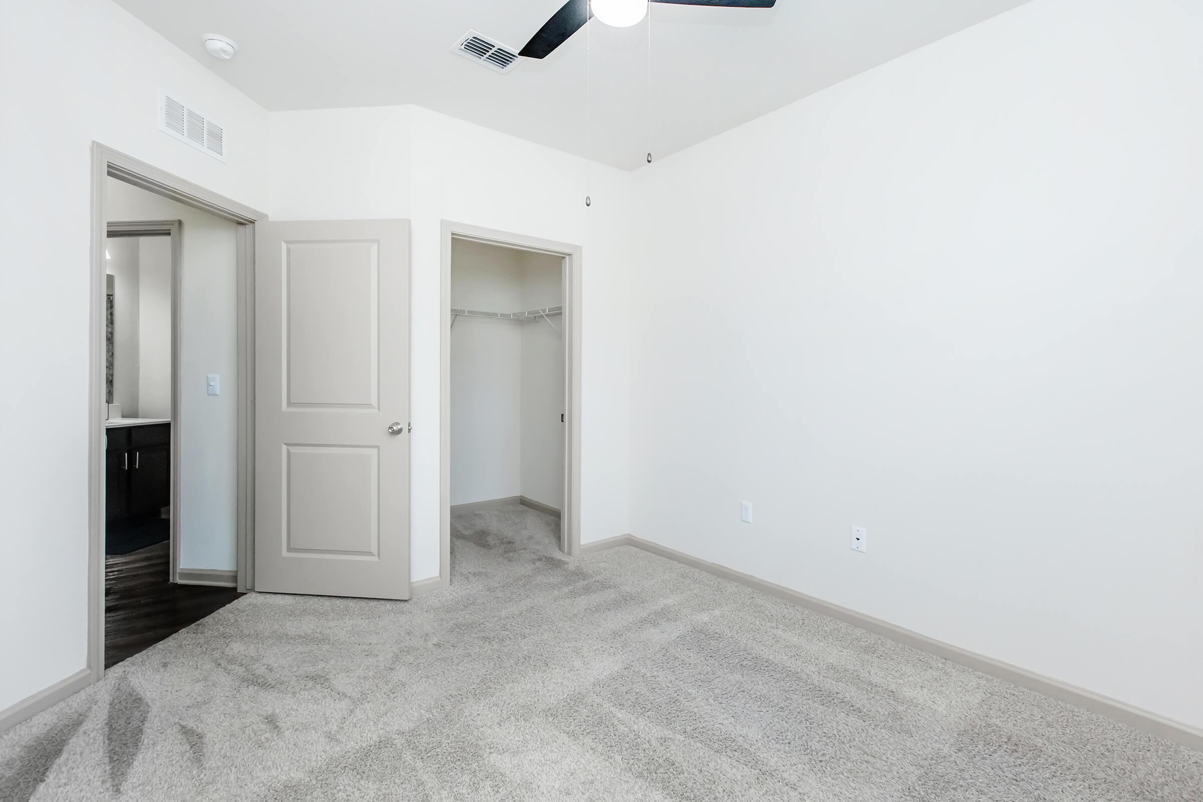Empty room with light-colored walls and plush carpet. There are two doorways leading to adjacent spaces, one of which appears to be a closet. A ceiling fan is mounted in the center of the room, and the overall atmosphere is bright and airy.