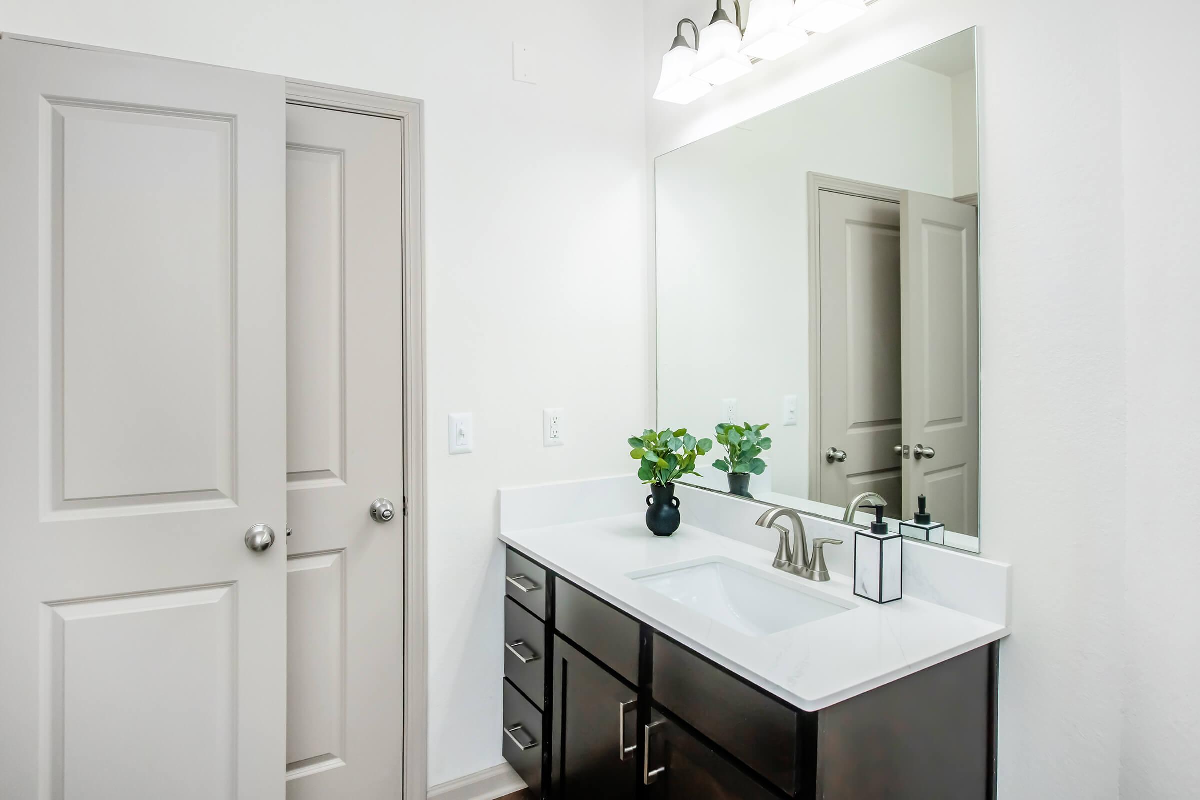 A modern bathroom featuring a white countertop with a sink, dark wooden cabinets, a mirror above the sink, and two doors partially visible. There are green plants on the counter and soft light from wall-mounted fixtures, creating a bright and clean atmosphere.