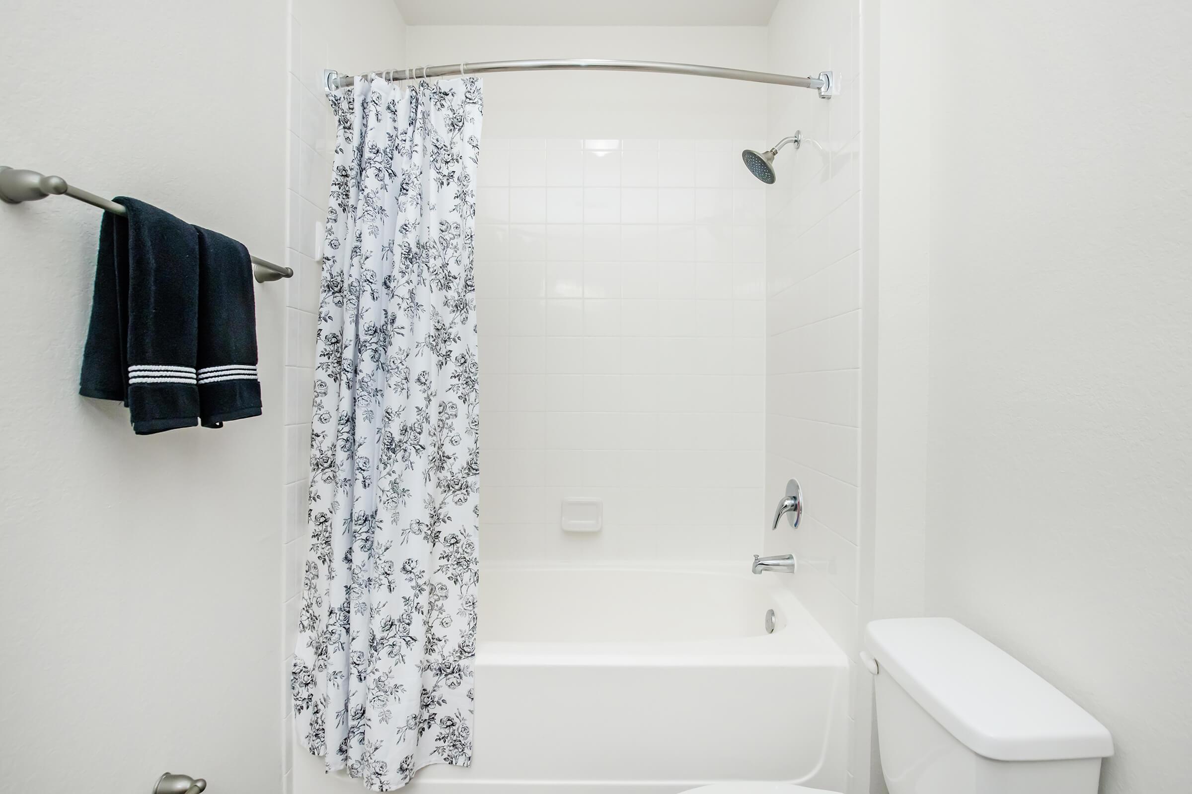 A clean bathroom setting featuring a white bathtub with a floral shower curtain, a handheld showerhead, and a toilet. A dark towel hangs on a rail beside the tub, and the walls and tiles are light-colored, creating a fresh and airy atmosphere.