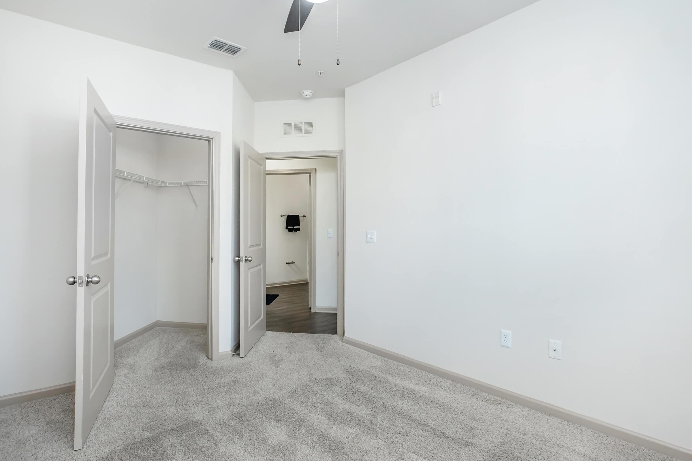 A clean, modern room with light-colored walls and carpet. There are two open doors: one leads to a closet, and the other to a bathroom. The bathroom is not fully visible. A ceiling fan is mounted above. The space is well-lit and minimalistic, giving it an airy feel.