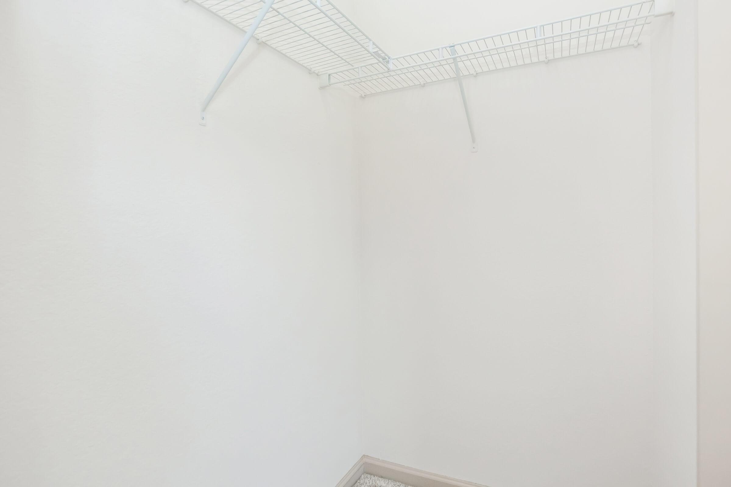 A clean, empty walk-in closet with white walls and a wire shelving unit mounted on the ceiling. The floor is partially covered with light-colored carpeting. The space has a bright, open feel, with ample lighting and no items or furniture present.