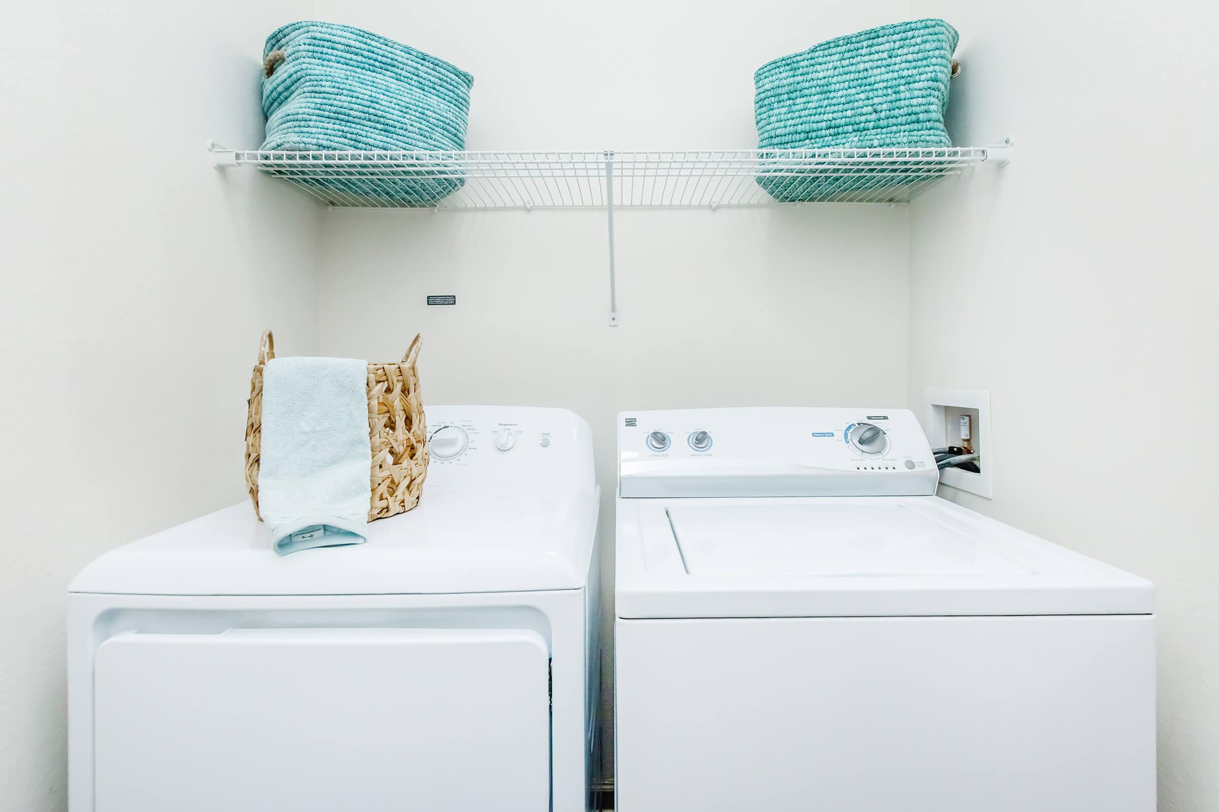 A laundry room featuring a washing machine and a dryer side by side. Above them, two woven baskets in shades of blue and a light blue towel are placed on a shelf. The walls are painted a light color, creating a clean and organized appearance.