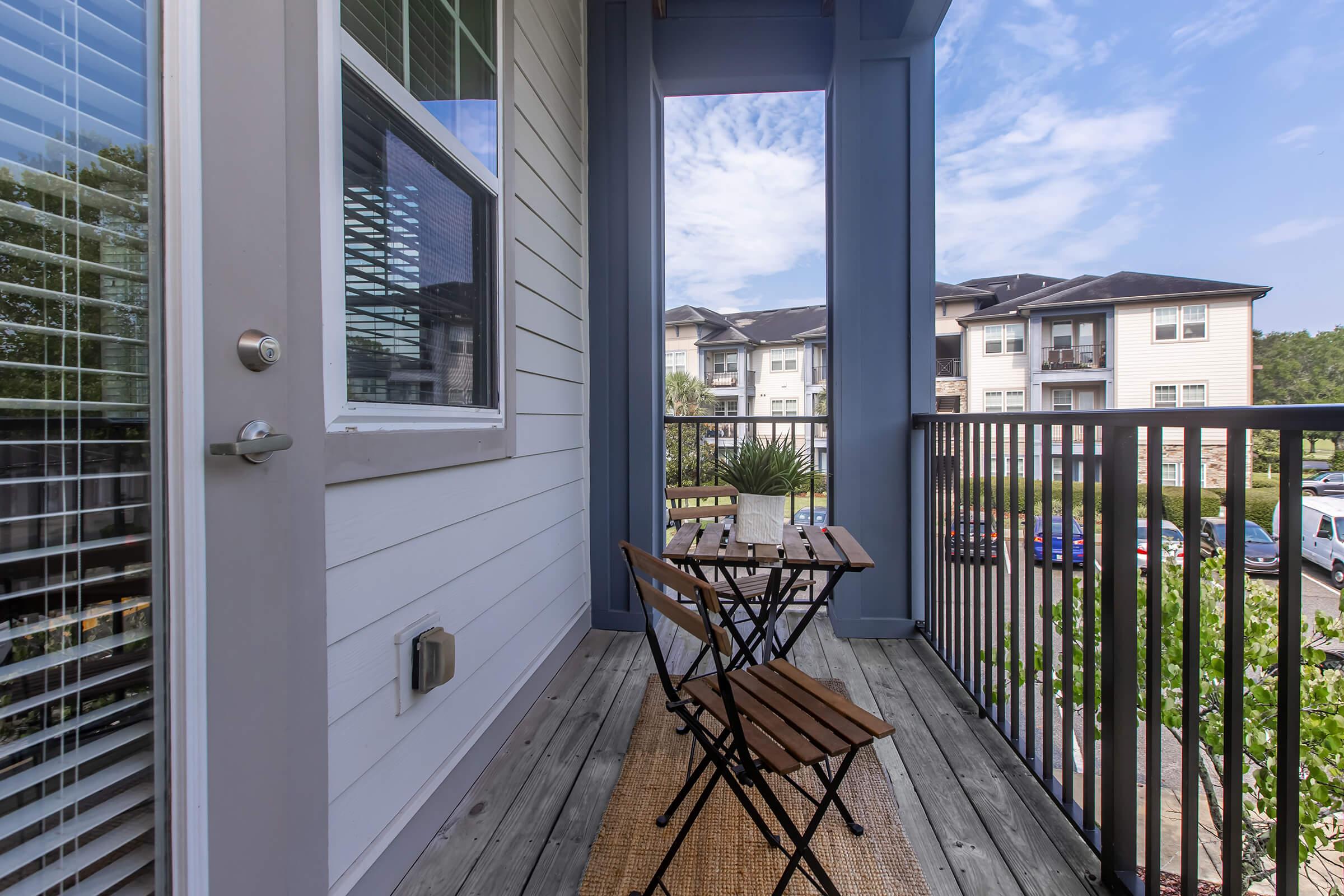 A small balcony with a wooden bistro table and two folding chairs, overlooking a view of apartment buildings and parked cars. The space features light-colored siding, large windows, and a wooden floor, with greenery visible in the background under a blue sky.