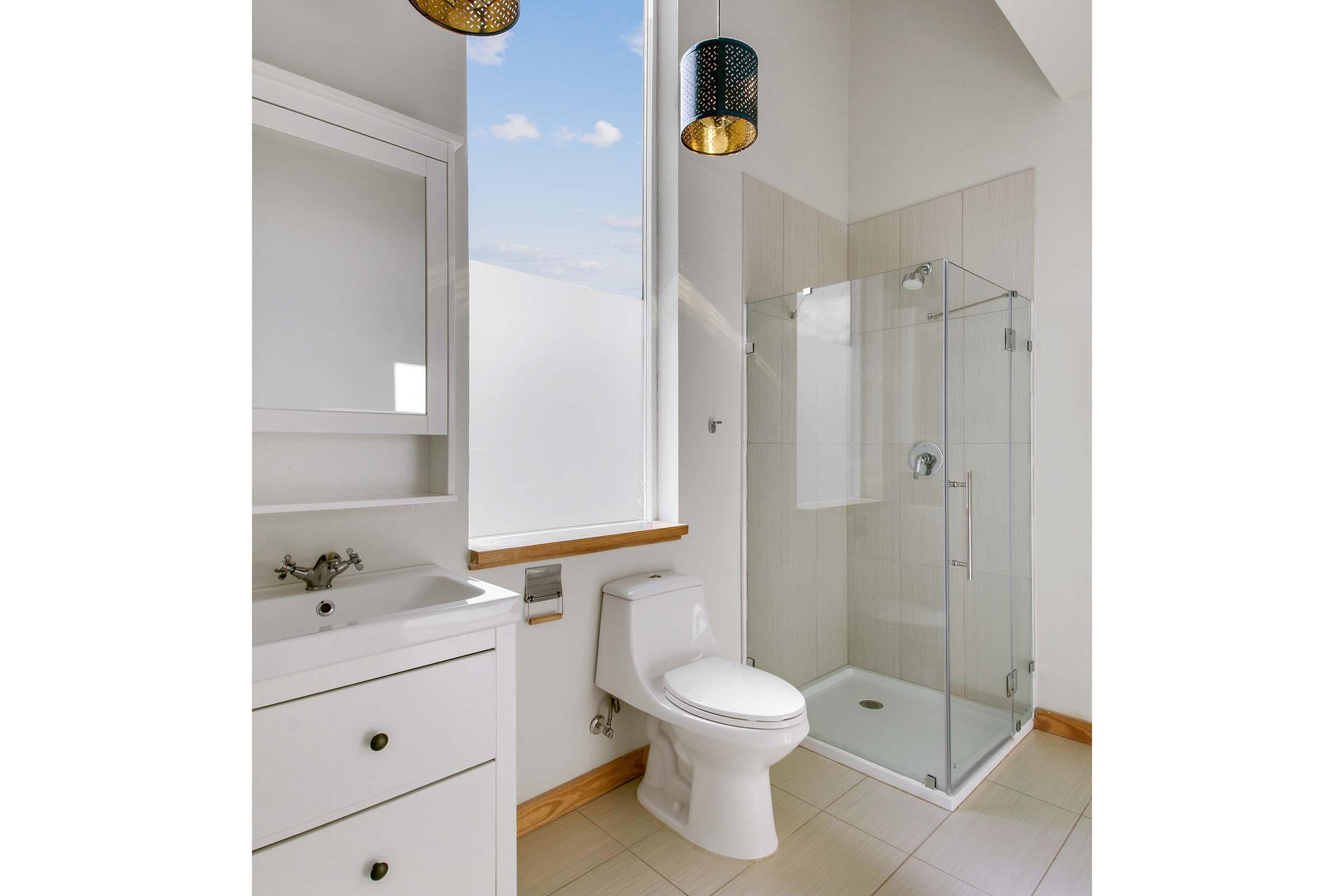 Modern bathroom featuring a glass shower enclosure, white toilet, and a vanity with a mirror. Natural light floods the space through large windows, highlighting the neutral color palette and clean lines. Stylish pendant lights hang from the ceiling.