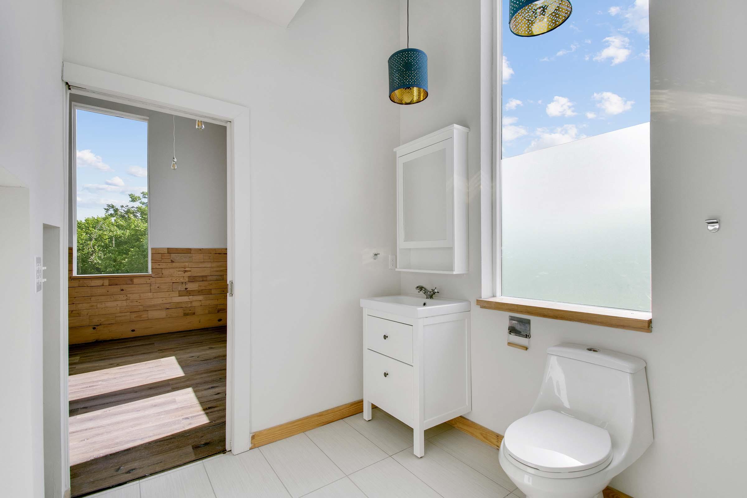 Bright and modern bathroom featuring a white toilet, a small vanity with a mirror, and large windows allowing natural light. The room has a neutral color scheme and a wooden accent wall visible through an open door, creating a spacious and airy atmosphere.