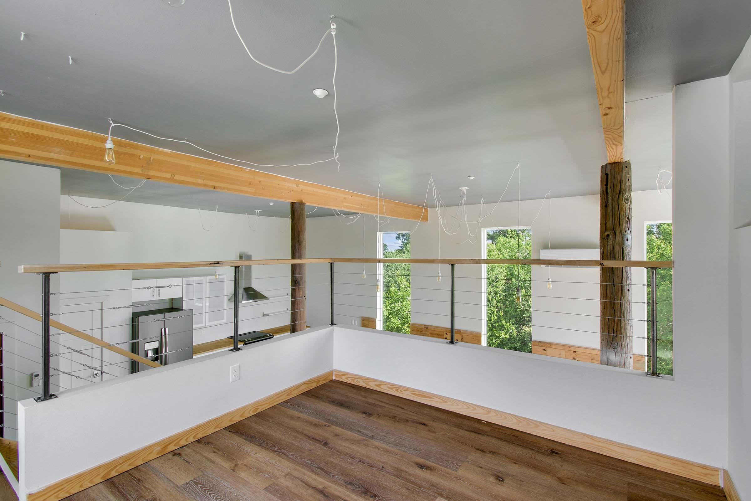 Interior view of a modern, open-concept living space featuring a wooden balcony with metal railings. The room has large windows allowing natural light to fill the space, and a minimalist design with unfinished ceiling light fixtures hanging down. Warm wooden flooring adds contrast to the white walls.