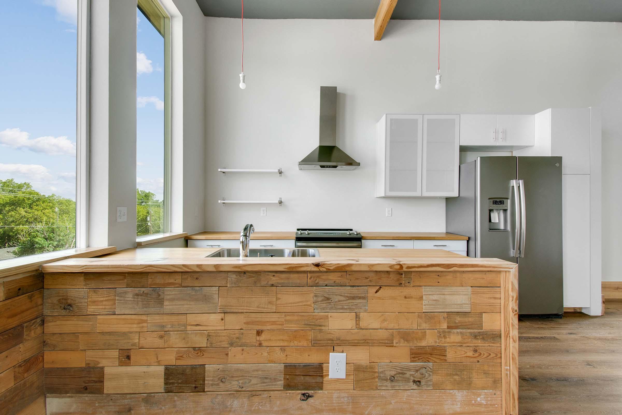A modern kitchen featuring a wooden bar with a tiled top, stainless steel appliances, and a large window that offers a view of greenery outside. The kitchen includes a refrigerator, stove, and sleek cabinetry. Bright natural light fills the space, highlighting the minimalist design.