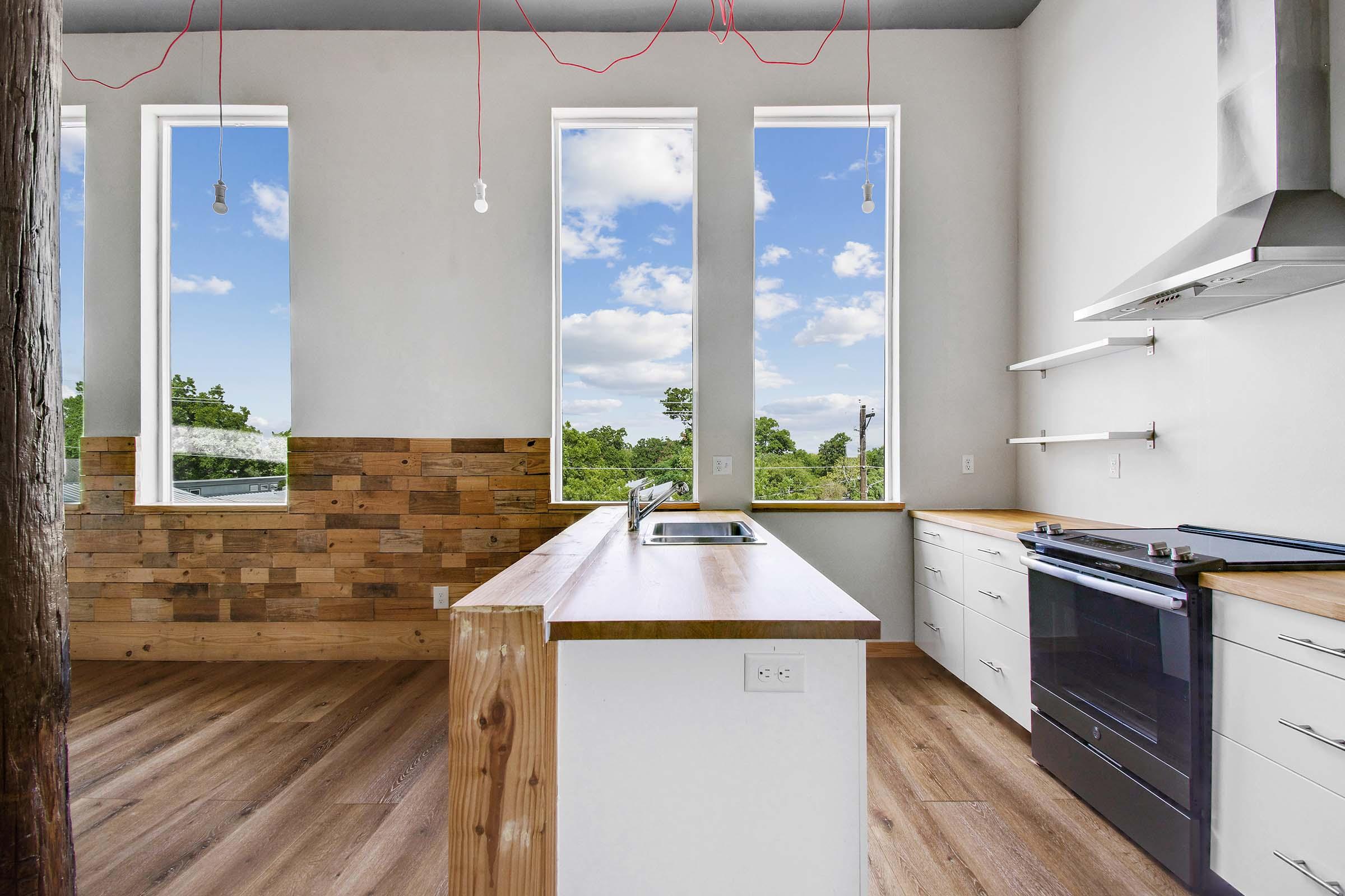 A modern kitchen with large windows offering a view of blue skies and greenery. The design features wooden accents, a central island with a sink, white cabinetry, and stainless steel appliances. The space has a minimalist aesthetic with natural light streaming in.