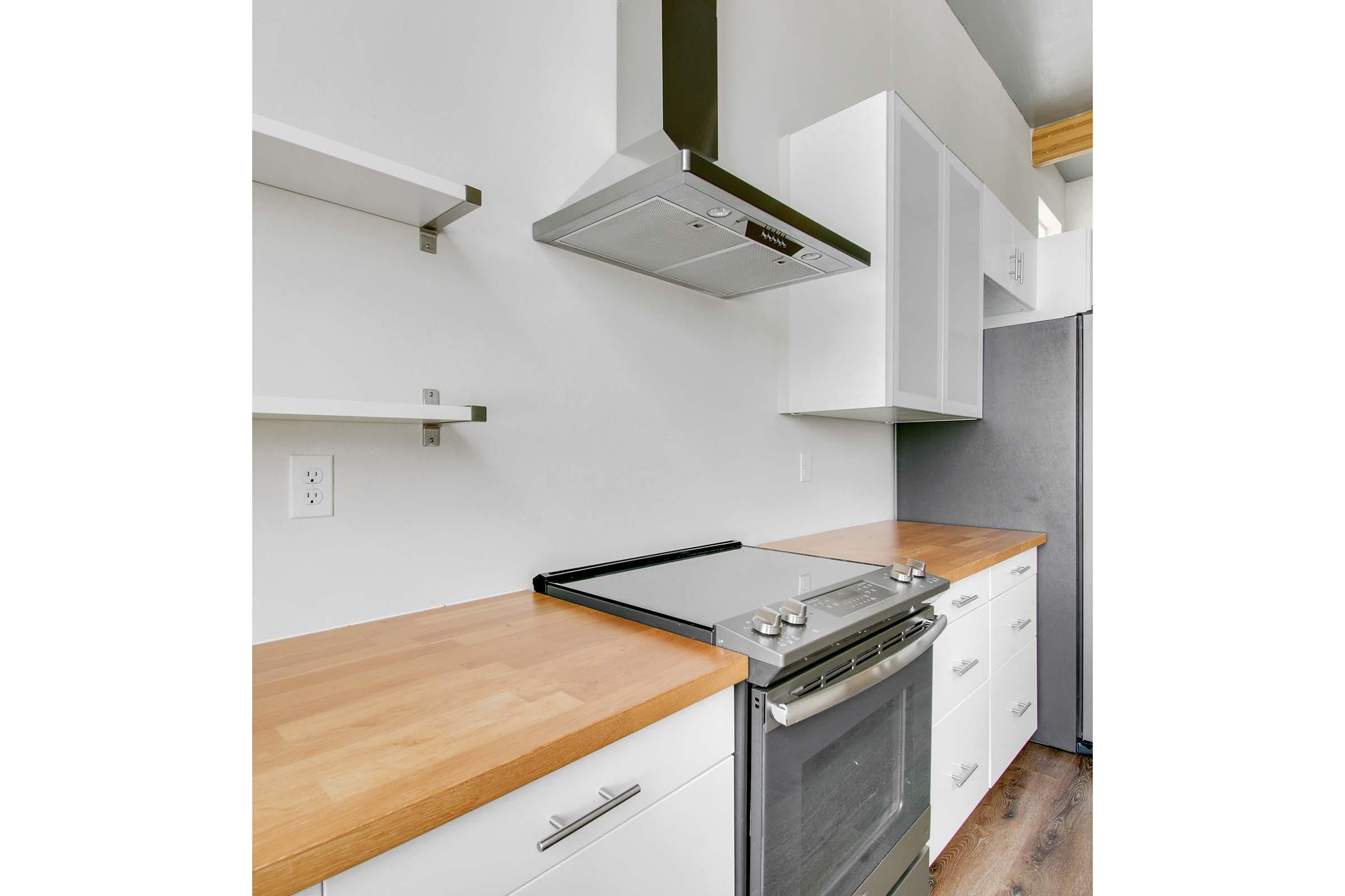 Modern kitchen featuring a stainless steel range with a sleek hood, wooden countertop, and minimalist white cabinets. Open shelving above the counter adds to the contemporary design. A refrigerator is visible in the corner, highlighting a clean and functional layout.