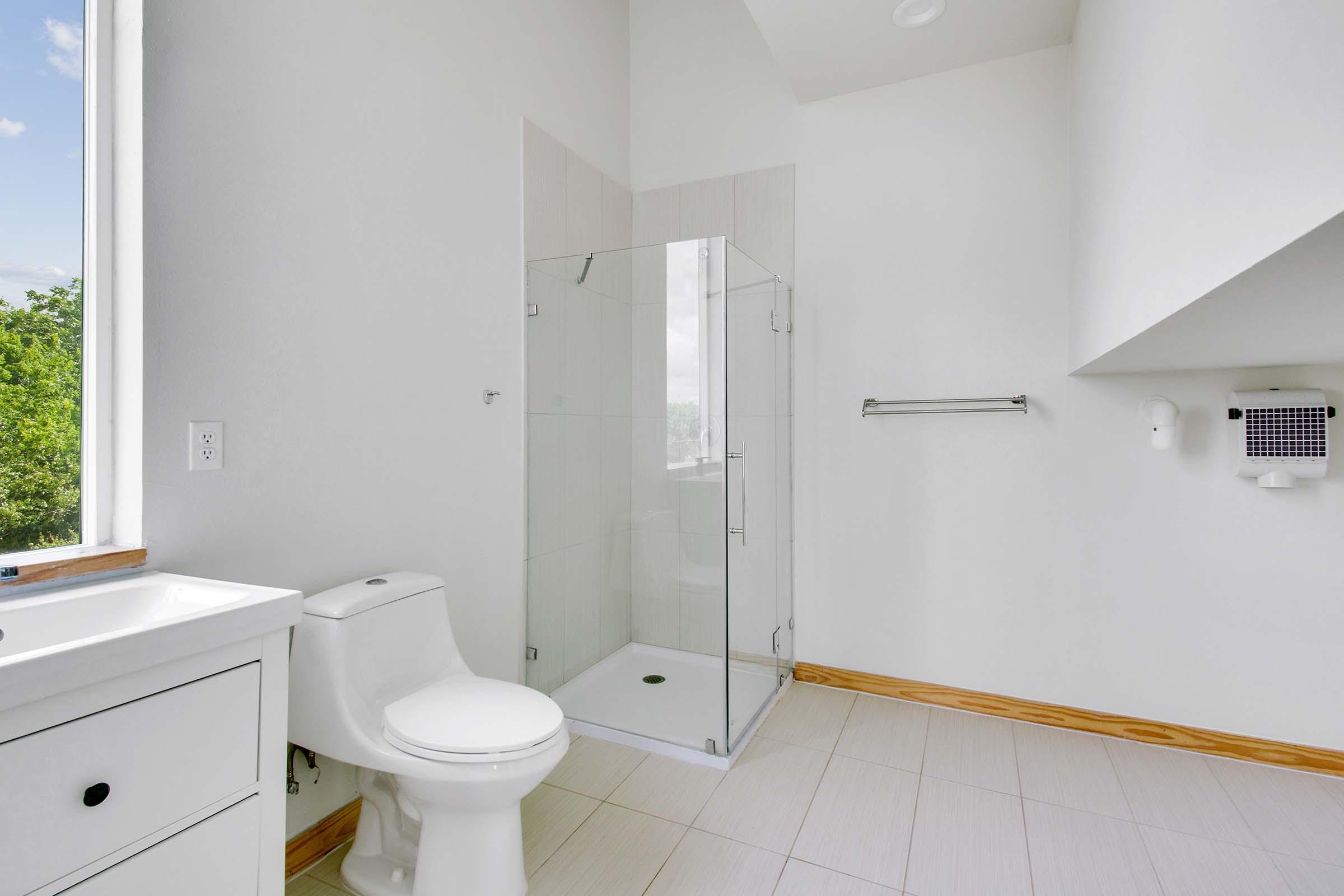 A modern bathroom featuring a white toilet, a glass-enclosed shower, and a white vanity with a mirror. Natural light streams in through a large window, illuminating the light-colored tiles and minimalistic design. A towel rack is installed on the wall, and an air hand dryer is mounted nearby.