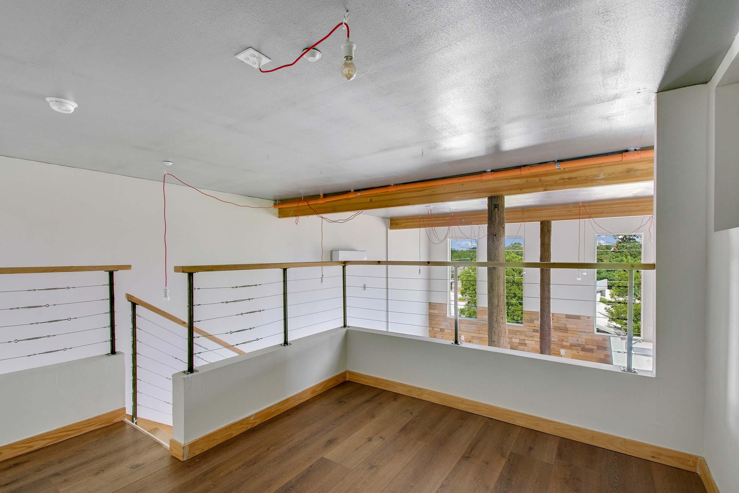 Interior view of a modern space with wooden flooring and a partial railing. The walls are painted white, and there are exposed electrical wires. Large windows allow natural light to fill the room, highlighting the minimalistic design. A bulb hangs from the ceiling, adding to the unfinished look of the area.