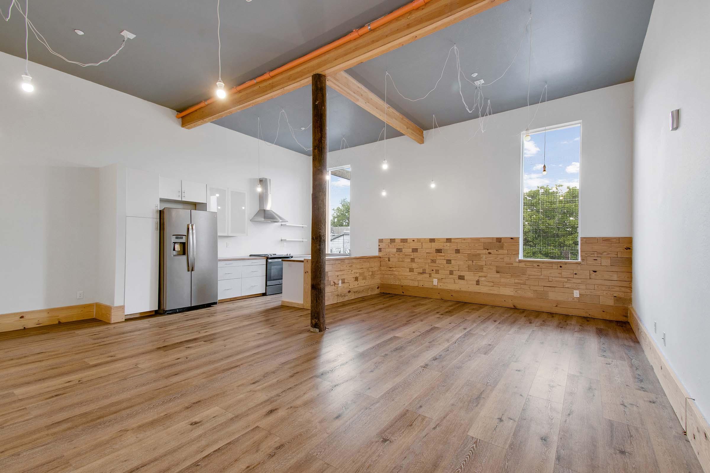A spacious, modern apartment interior featuring wooden flooring, a partially wooden wall, and a kitchen area with stainless steel appliances. The ceiling is adorned with hanging lights, and large windows provide natural light, enhancing the bright and airy atmosphere.