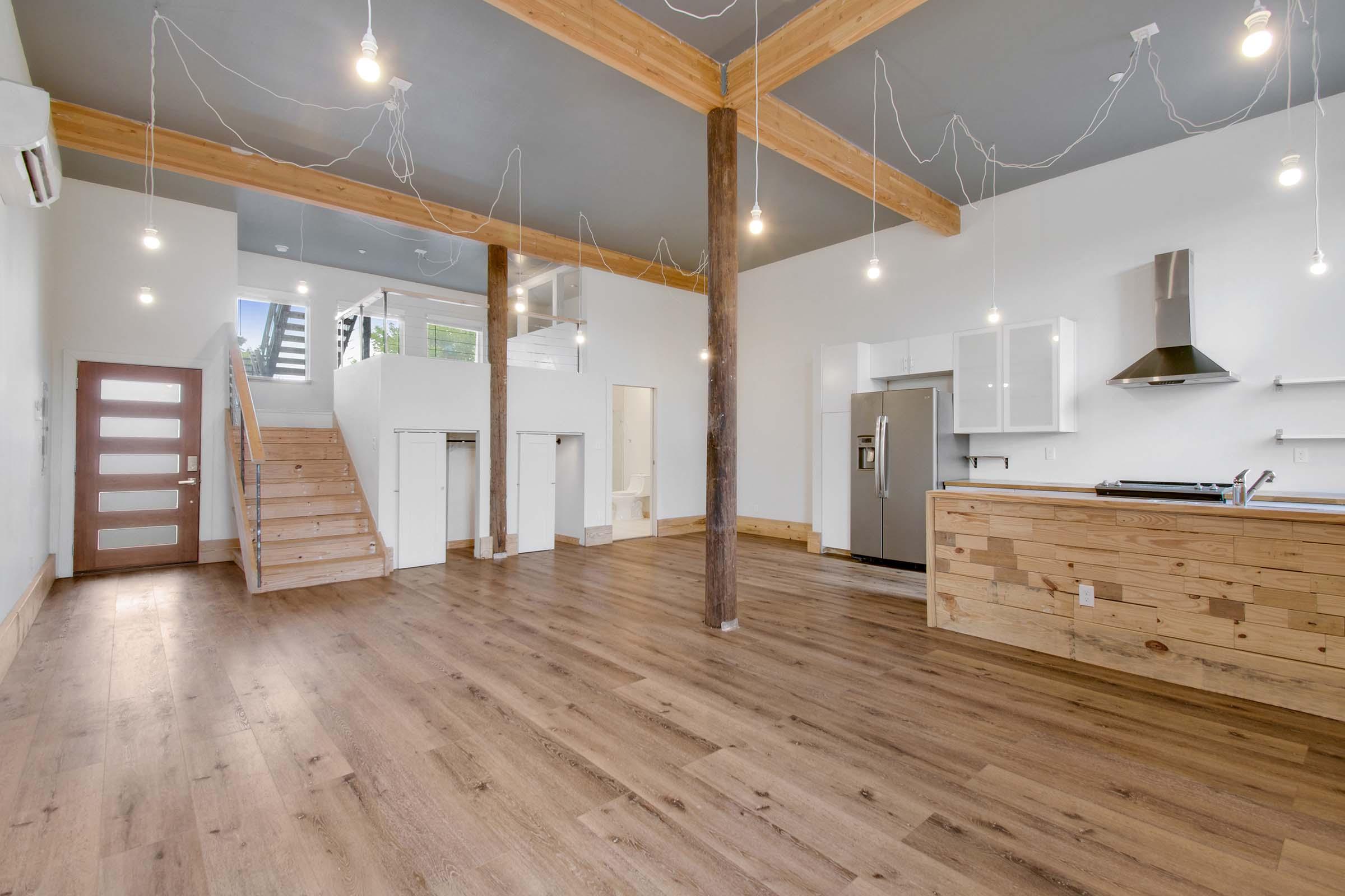 Spacious modern interior featuring an open-plan living area with wooden floors, large windows, and stylish fixtures. The kitchen has a central island and stainless-steel appliances. A staircase leads to a second level, with minimalist design elements and bright lighting from hanging bulbs.