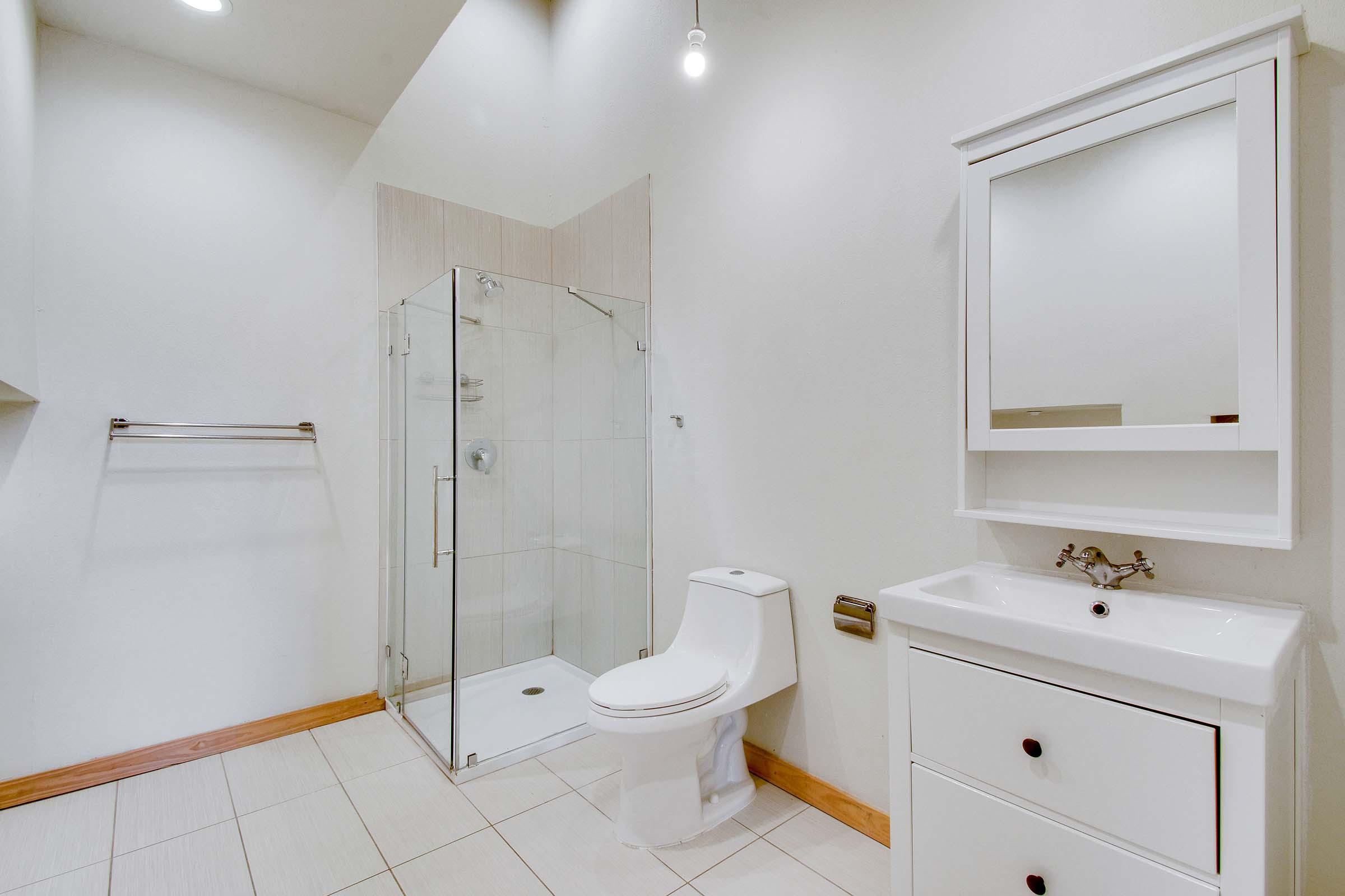 A modern bathroom featuring a glass shower enclosure, a toilet, and a sink with a cabinet below. The walls are painted a light color, and there is a mirror above the sink. The flooring is tiled, and there are minimal decorative elements, creating a clean and open space.