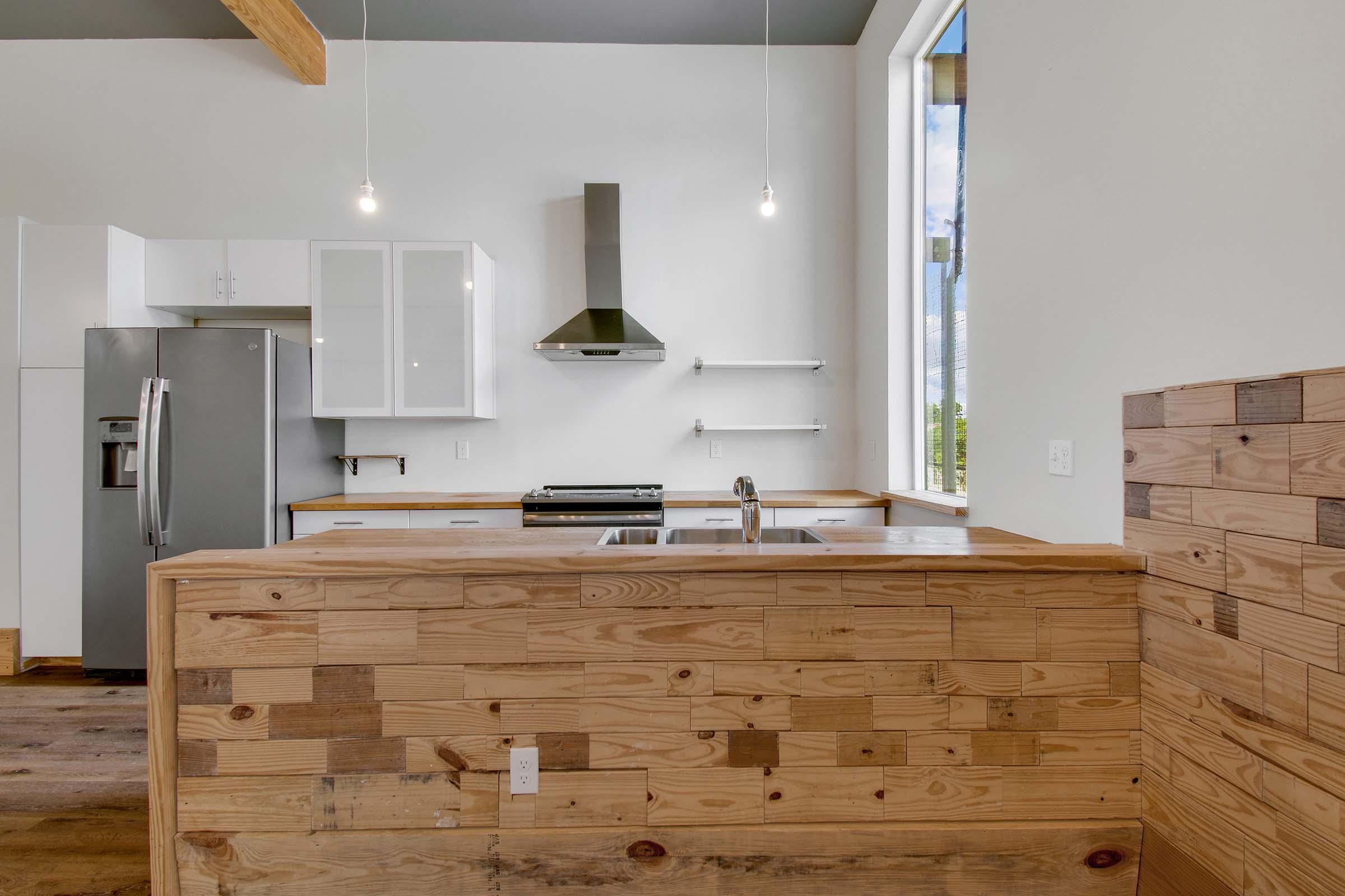 A modern kitchen featuring wooden cabinetry, a stainless steel refrigerator, and a sleek stove. The design includes an open layout with large windows allowing natural light, complemented by minimalist shelves and pendant lighting hanging from the ceiling.