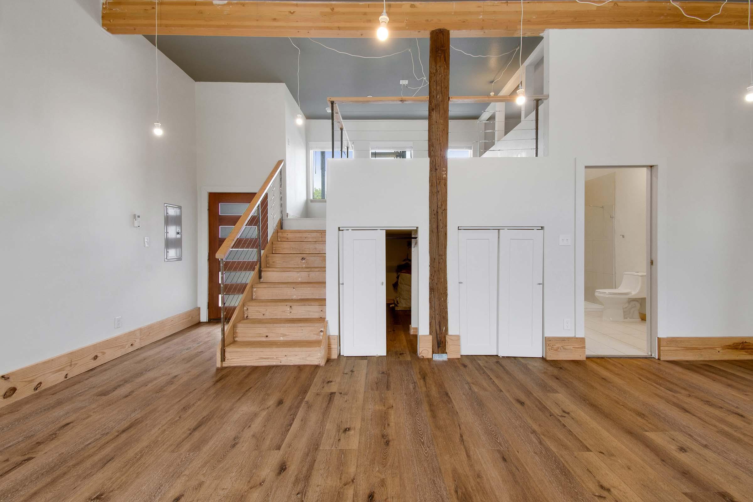 Interior of a modern, minimalist living space featuring wooden beams and flooring. There is a staircase to the left, leading to an upper level. On the right, a small bathroom door is visible. Closets are built into the wall, and the area is well-lit with pendant lights. The walls are painted white, creating an open, airy feel.