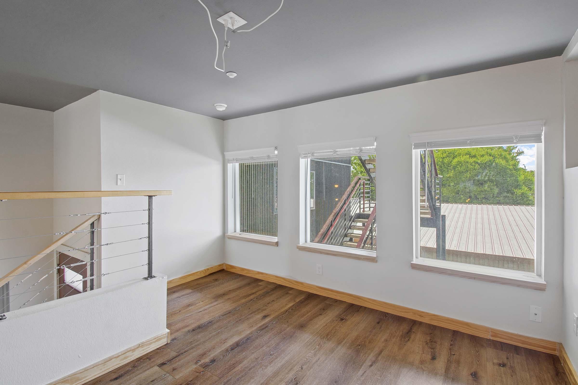 Bright, spacious room with a modern design, featuring three large windows allowing plenty of natural light. The walls are painted a soft color, and the flooring is a warm wood finish. A railing can be seen leading to a stairway in the background, adding to the open feel of the space.