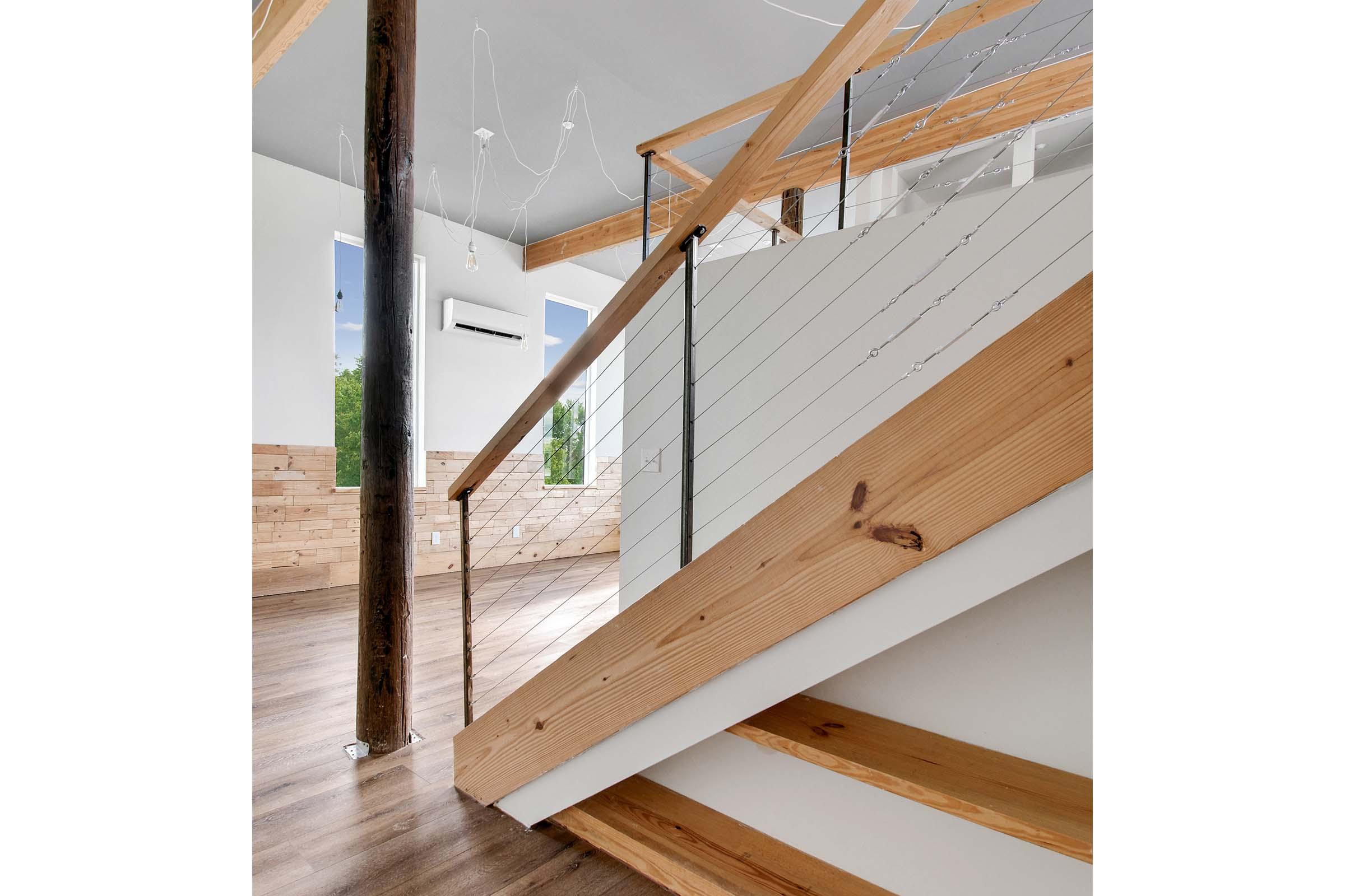 Interior view of a modern staircase featuring wooden stairs, a minimalistic railing, and large windows allowing natural light to enter. The space has a contemporary design with exposed wooden beams and a clean, open aesthetic. The flooring is light wood, contributing to a warm ambiance.