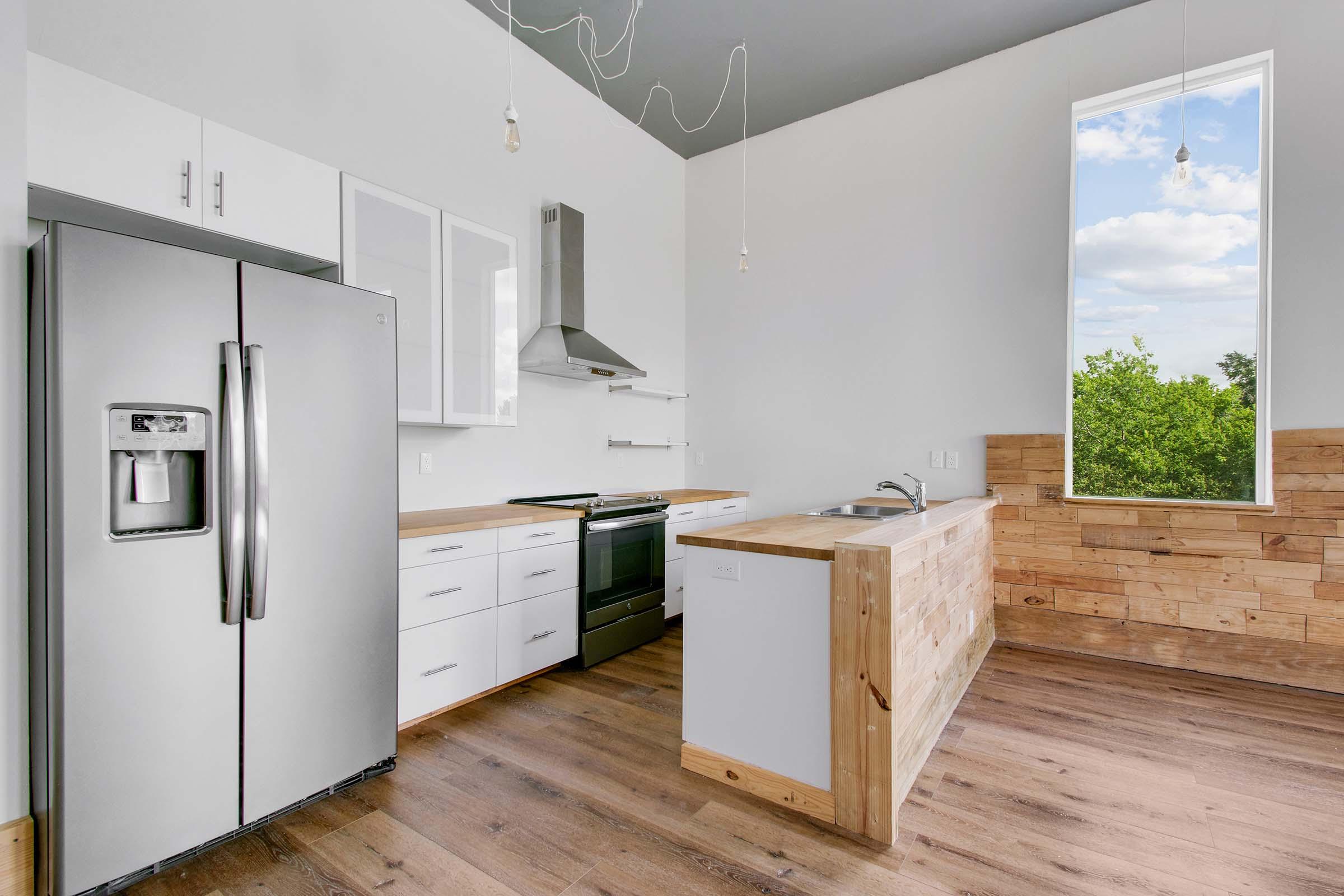 Modern kitchen with stainless steel appliances including a refrigerator and oven. Features white cabinets, a wooden island, and a large window providing natural light and a view of greenery outside. The flooring is wooden, complementing the contemporary design.