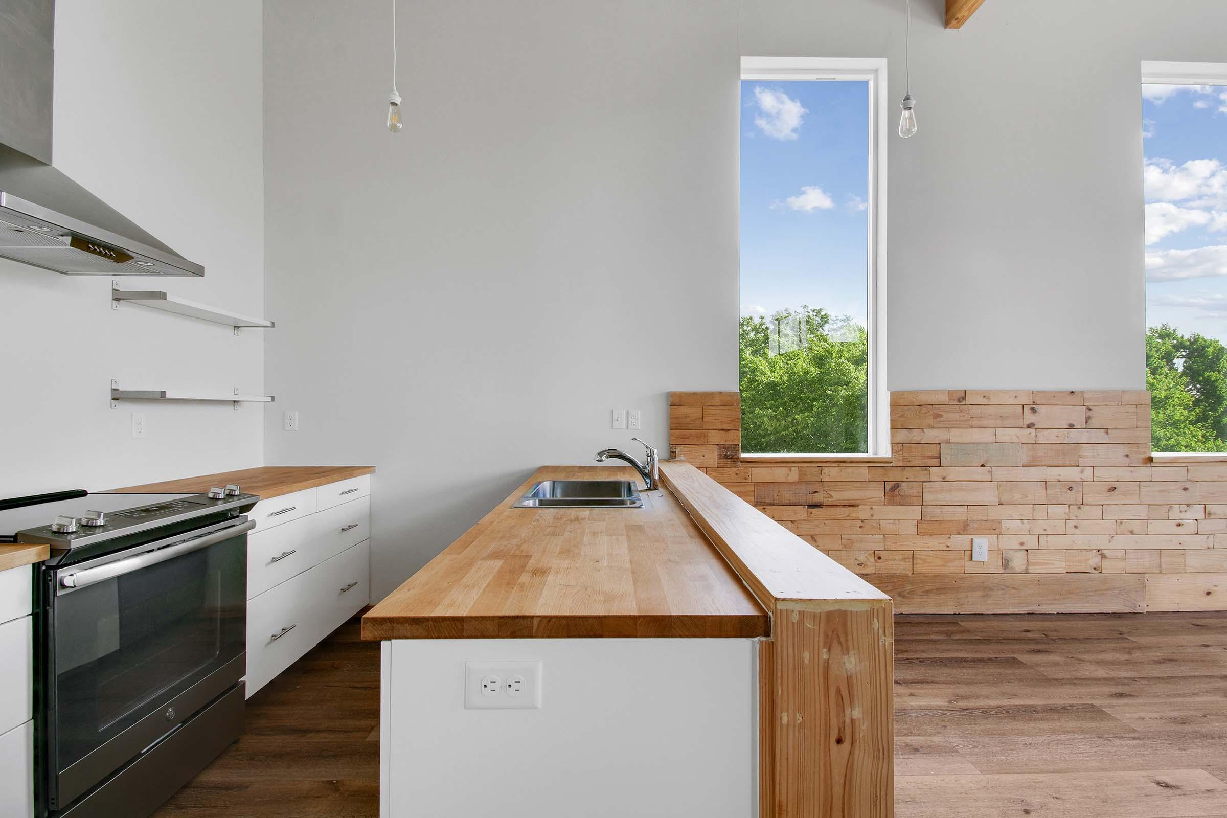 A modern kitchen featuring a wooden countertop, stainless steel oven, and white cabinetry. Large windows allow natural light to flood the space, showcasing greenery outside. The walls have a mix of light paint and wood paneling, creating a warm, contemporary atmosphere.