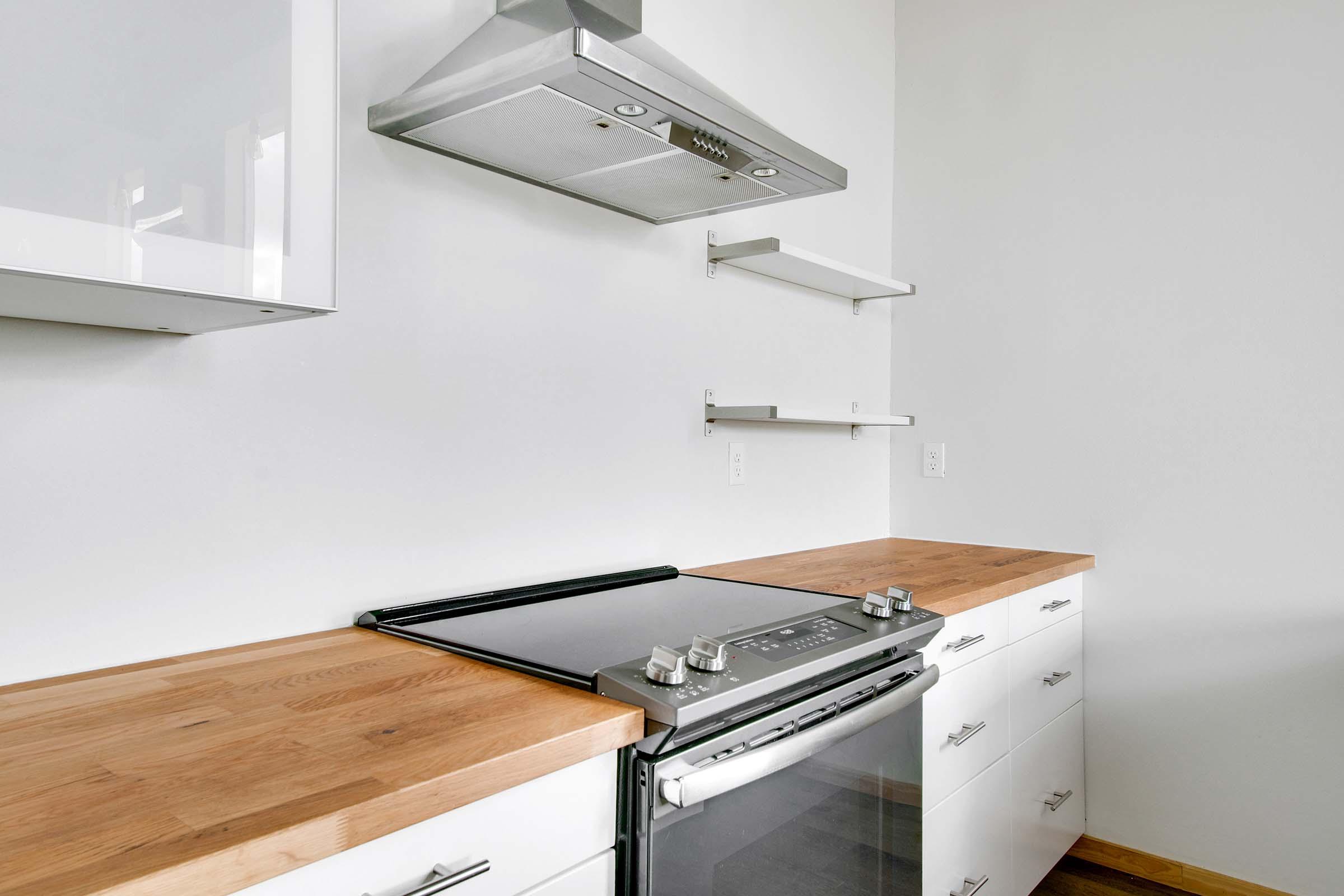 Modern kitchen featuring a sleek stove with an oven below, a stainless steel range hood, and light wooden countertops. White cabinets and open shelves provide a clean and minimalist aesthetic against a neutral wall.