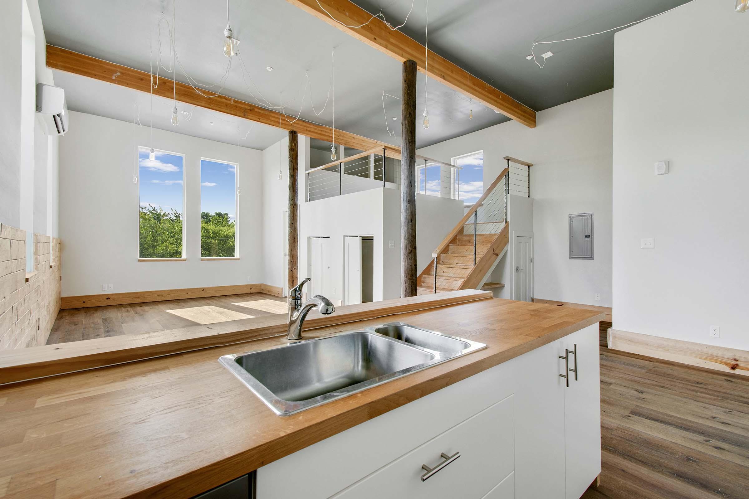 Bright, open living space featuring wooden beams and a modern kitchen with a stainless steel sink. Large windows allow natural light to fill the room, highlighting the wooden flooring. A staircase leads to an upper level, while minimalistic design elements create a clean and airy atmosphere.